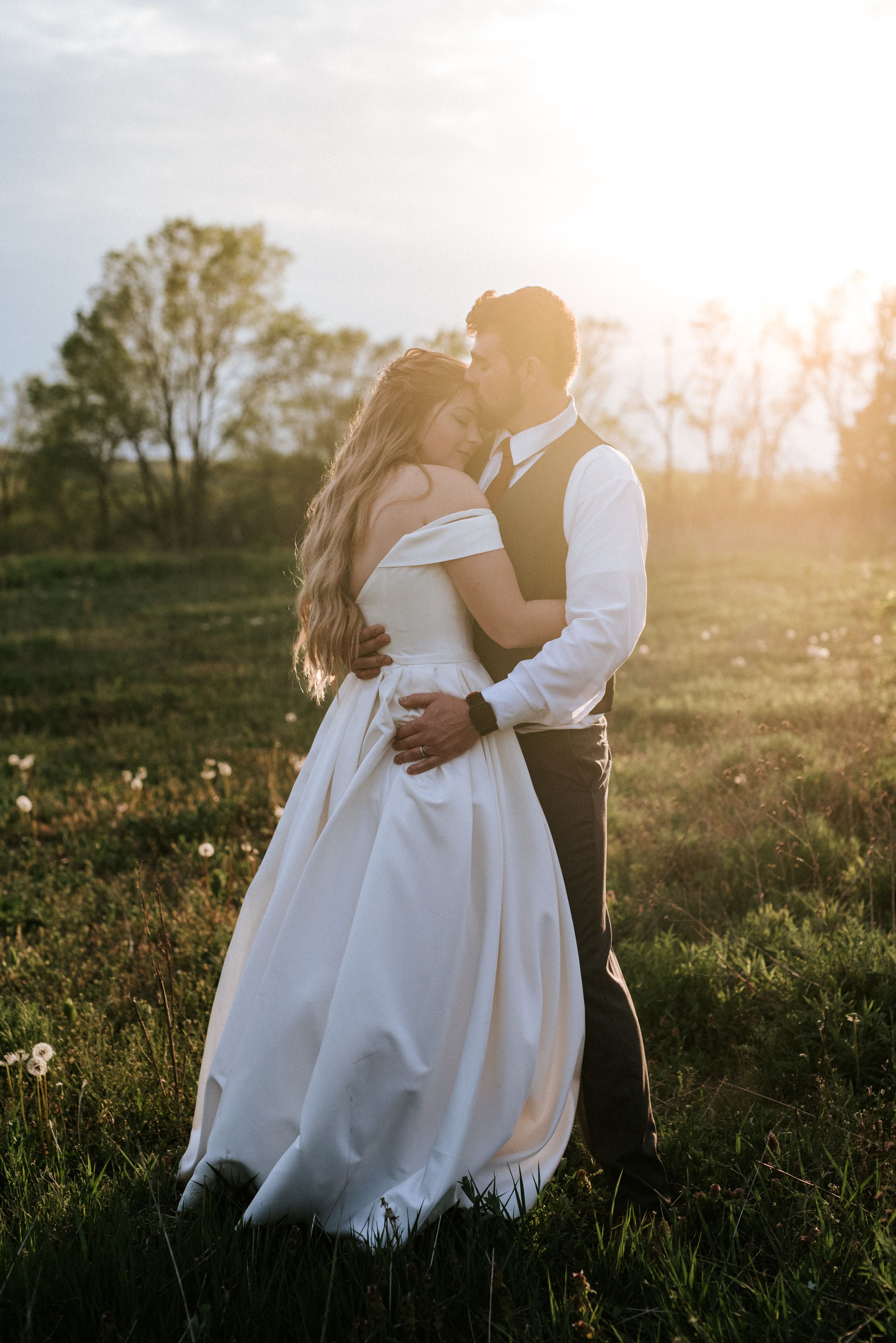Newlyweds embrace in golden sunlight during sunset in a rustic outdoor field setting.