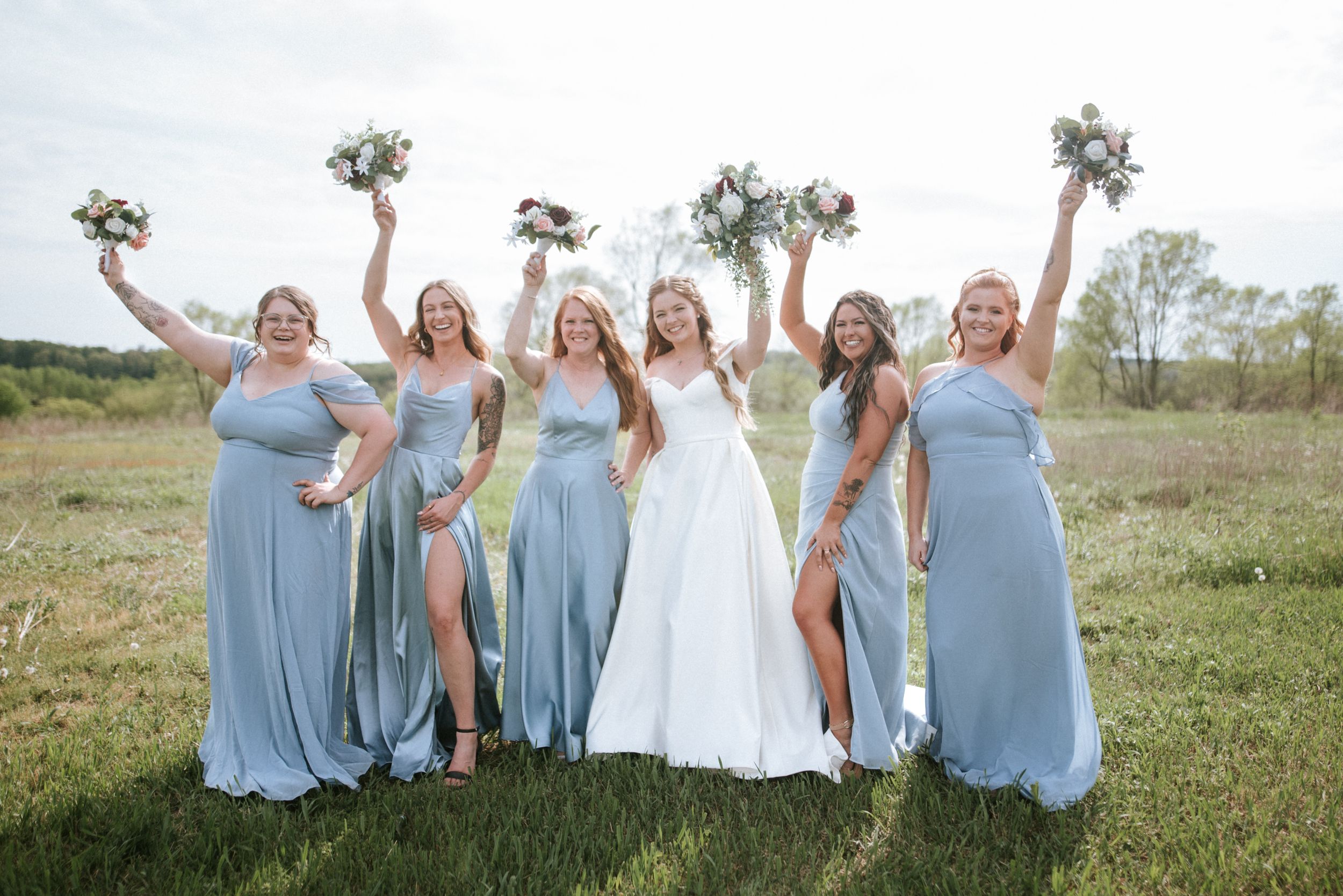 Bridesmaids in light blue dresses holding bouquets pose with bride in outdoor setting.