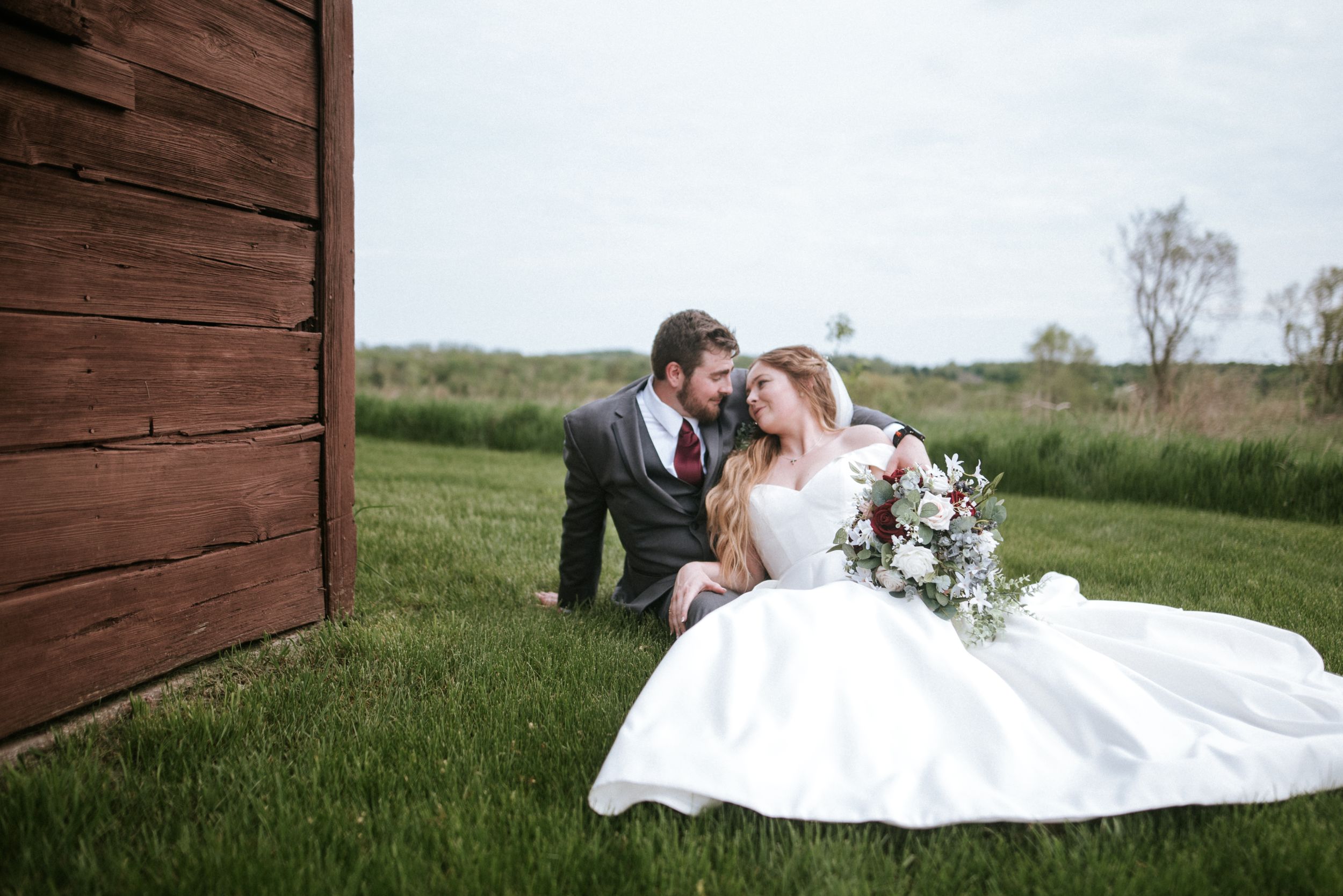 Newlyweds share a tender moment on grass beside a rustic wooden barn.