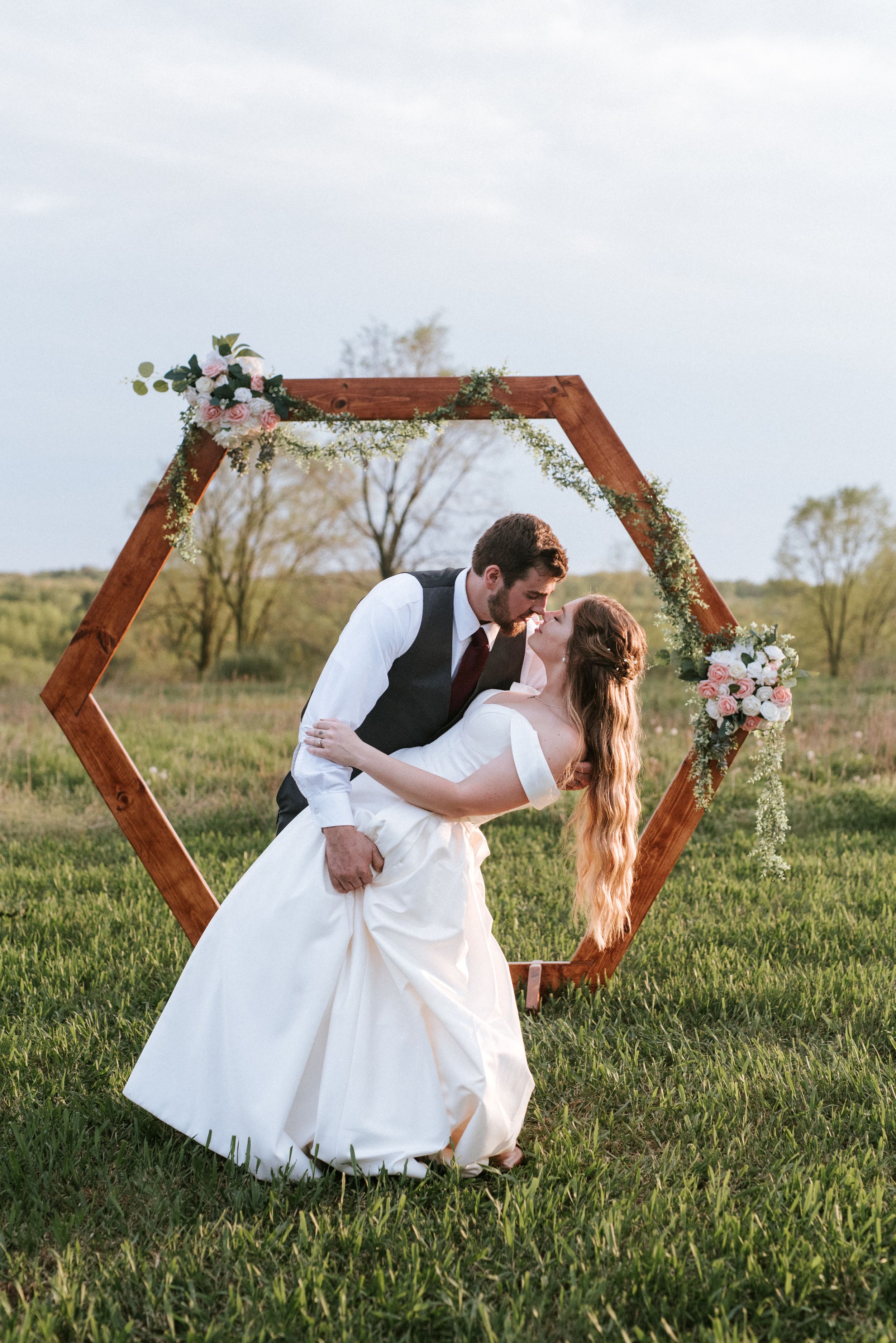 Romantic wedding photo featuring a wooden hexagon arch decorated with flowers as couple shares intimate moment in field.