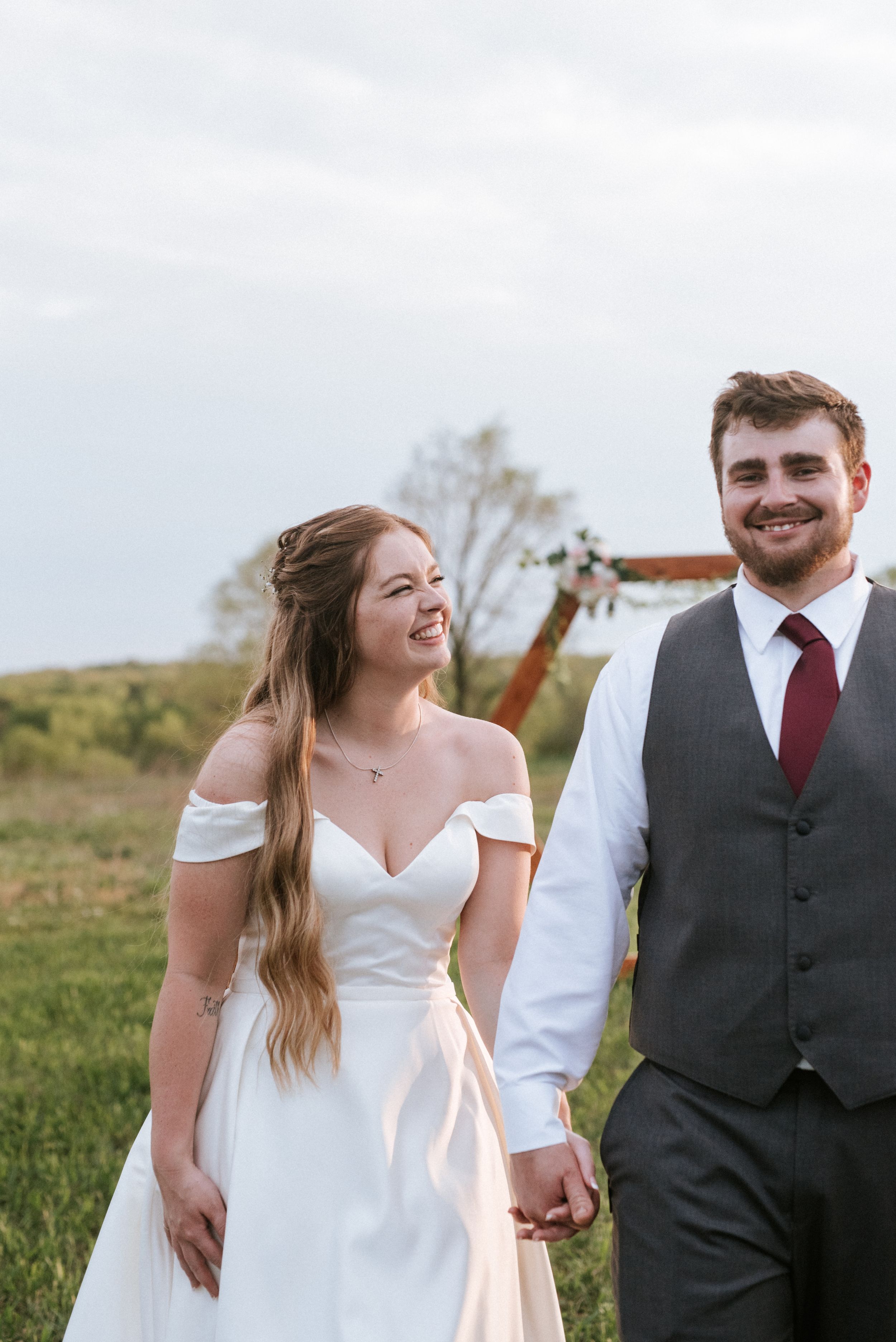 Wedding couple in rustic outdoor setting with bride in off-shoulder dress and groom in grey vest.