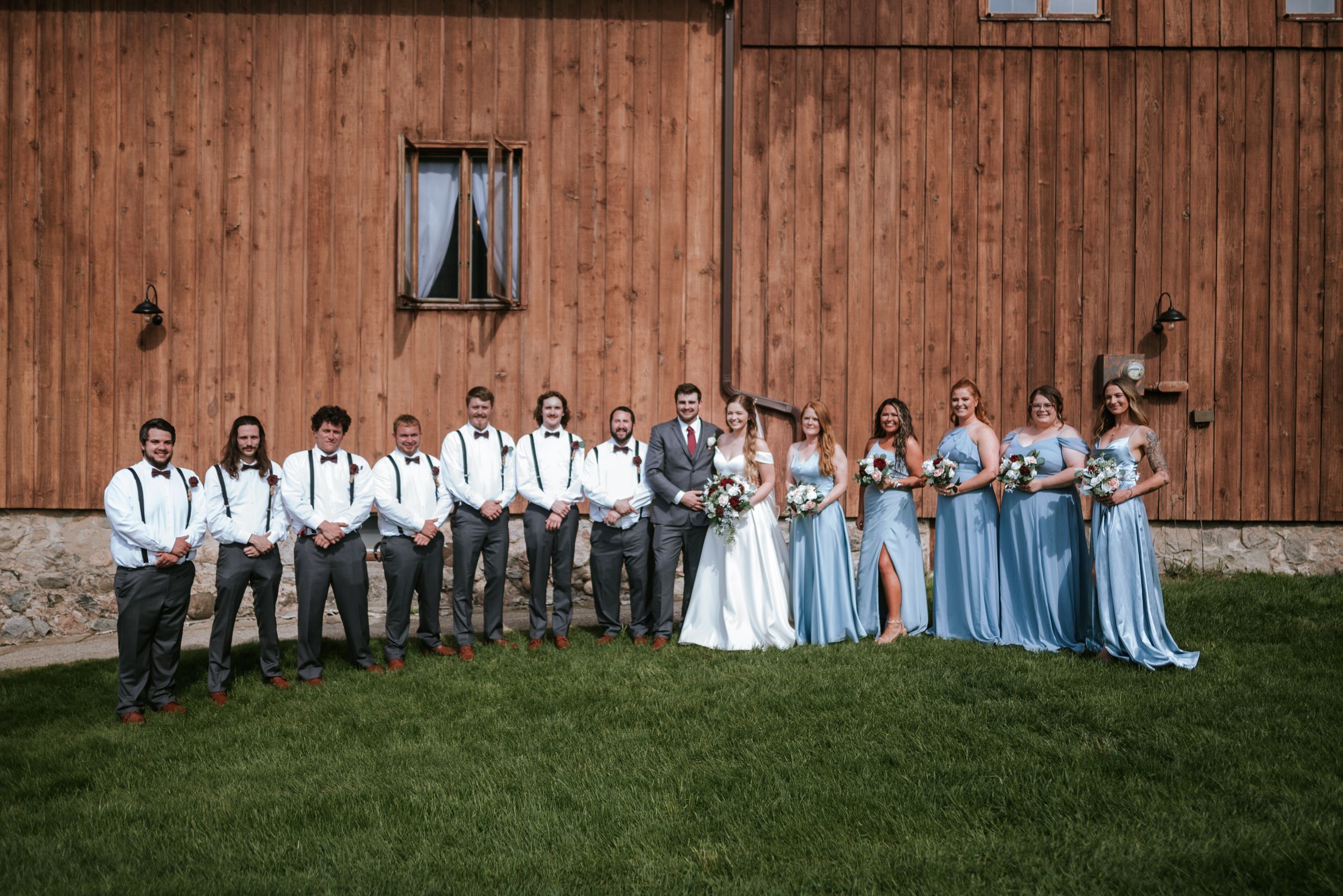 Wedding party in blue and grey attire poses together in front of rustic wooden barn.