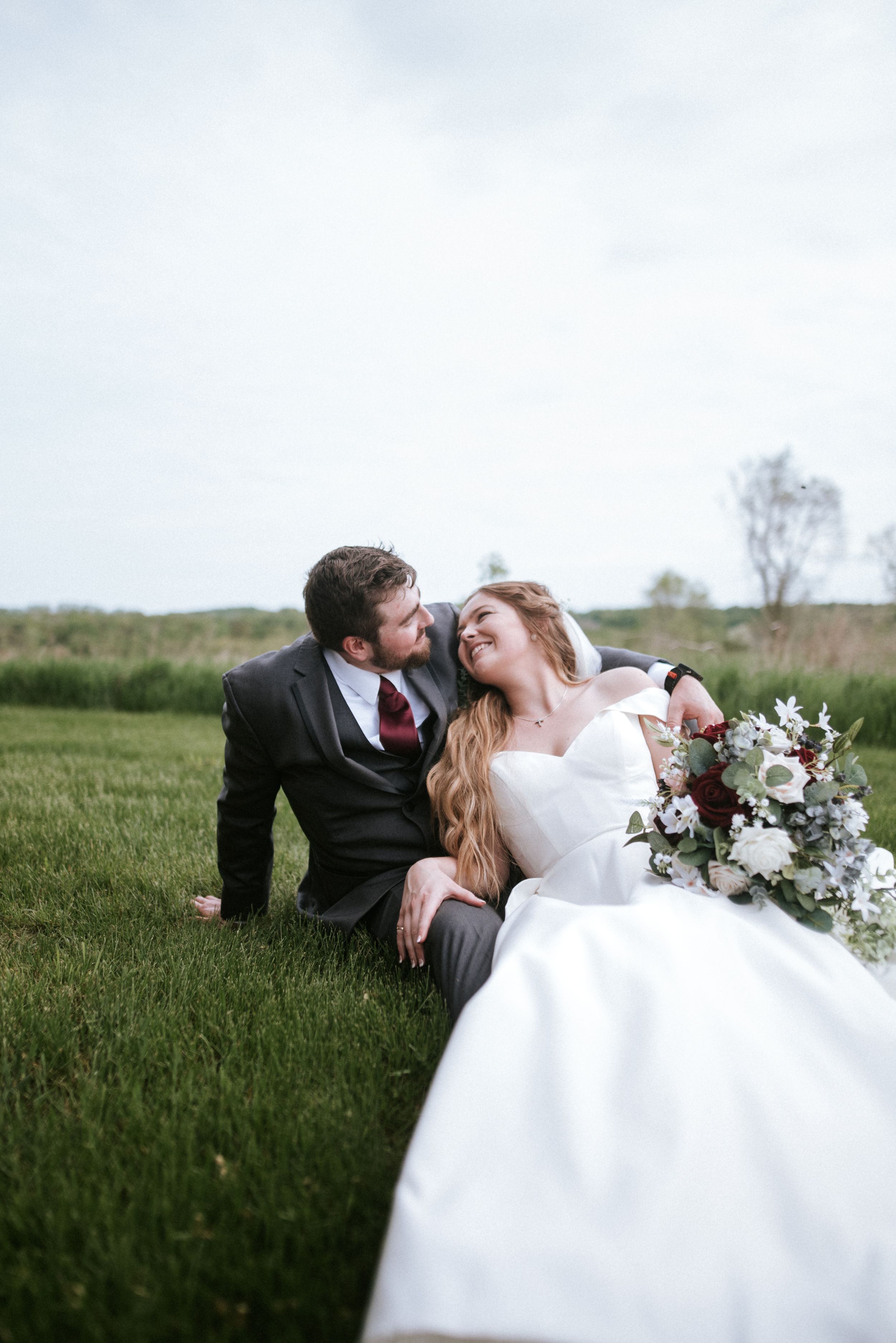 A romantic wedding portrait of a couple relaxing in a grassy field with a beautiful bouquet.