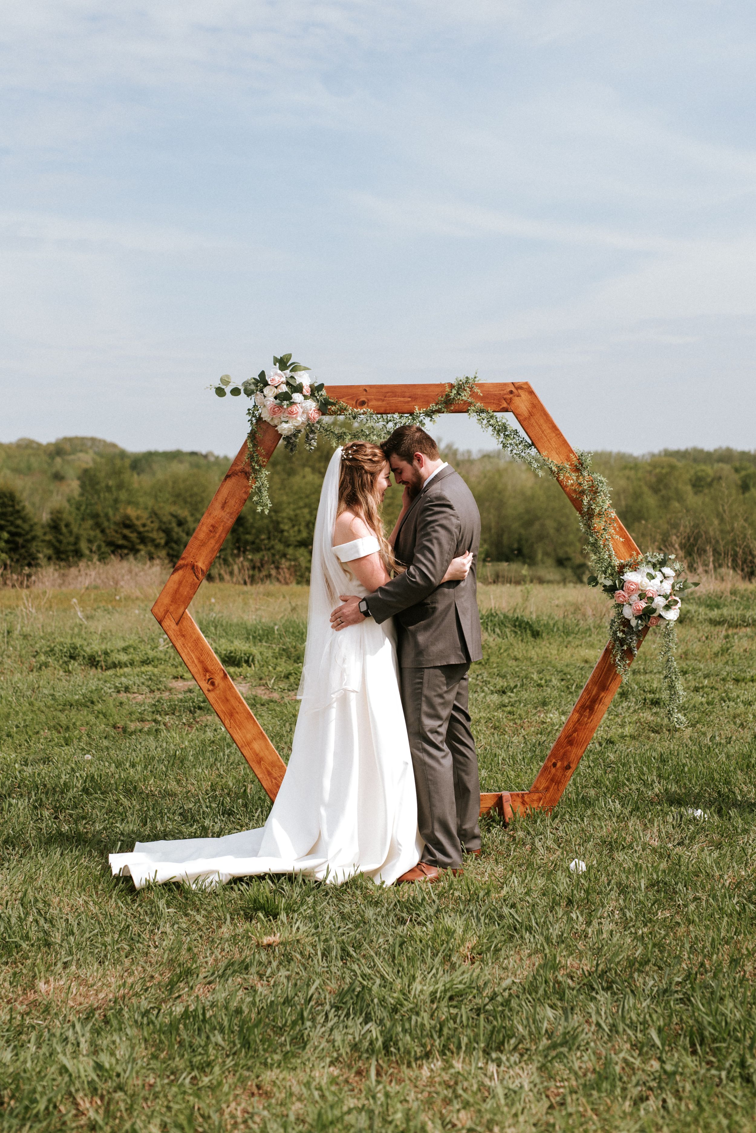 Couple shares intimate moment under hexagonal wooden wedding arch decorated with white flowers in field.