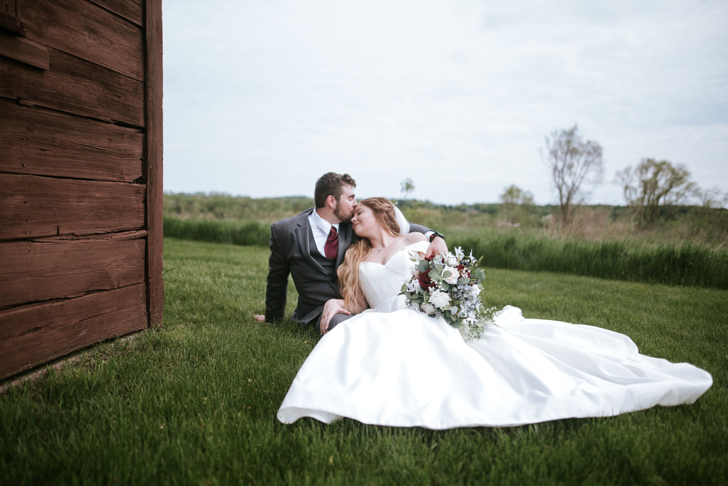 Outdoor wedding portrait of couple sitting on grass by rustic red barn wall.