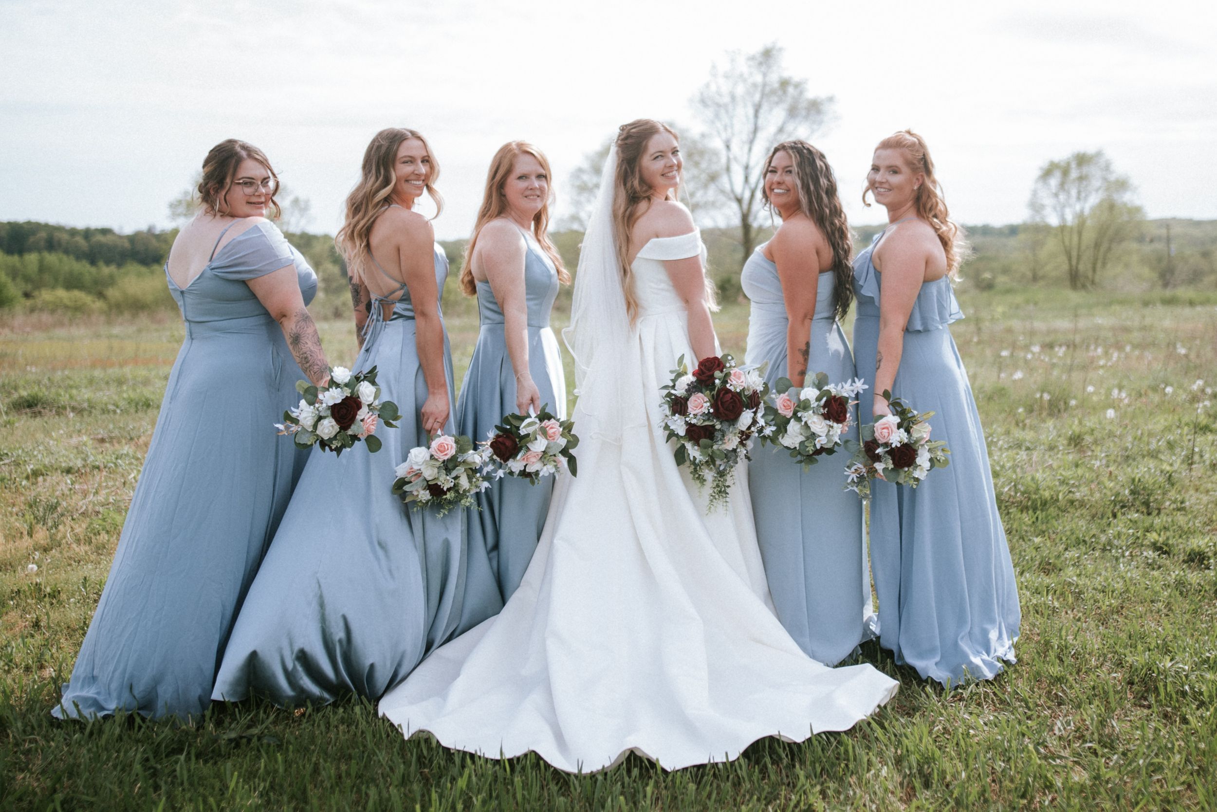 Bride and bridesmaids in flowing blue dresses pose with bouquets in countryside setting.