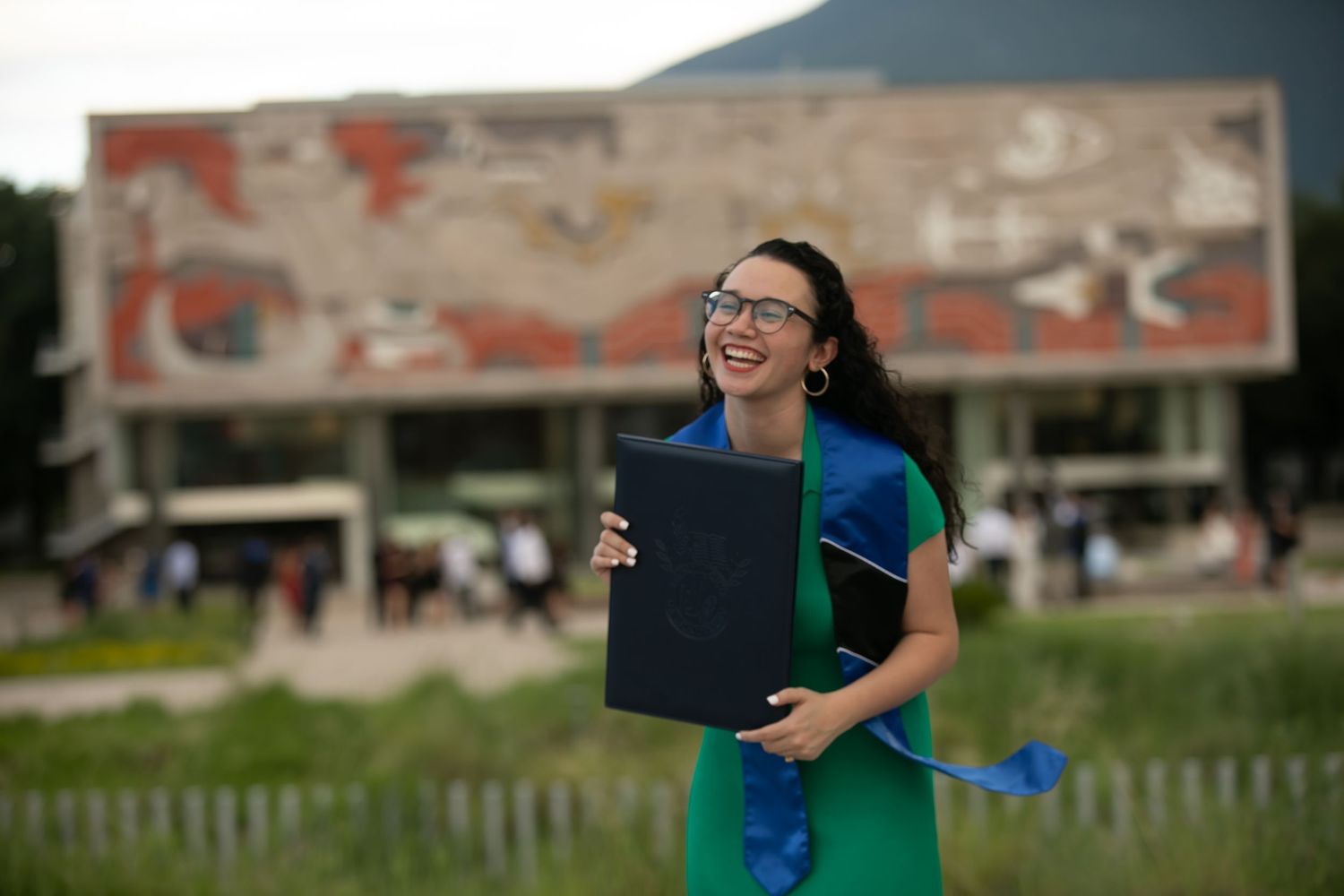 Happy woman in green dress holding diploma folder outside university campus building.