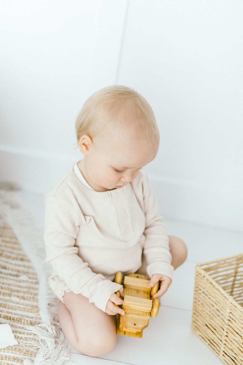 Baby in cream colored outfit playing with wooden toy on textured rug.