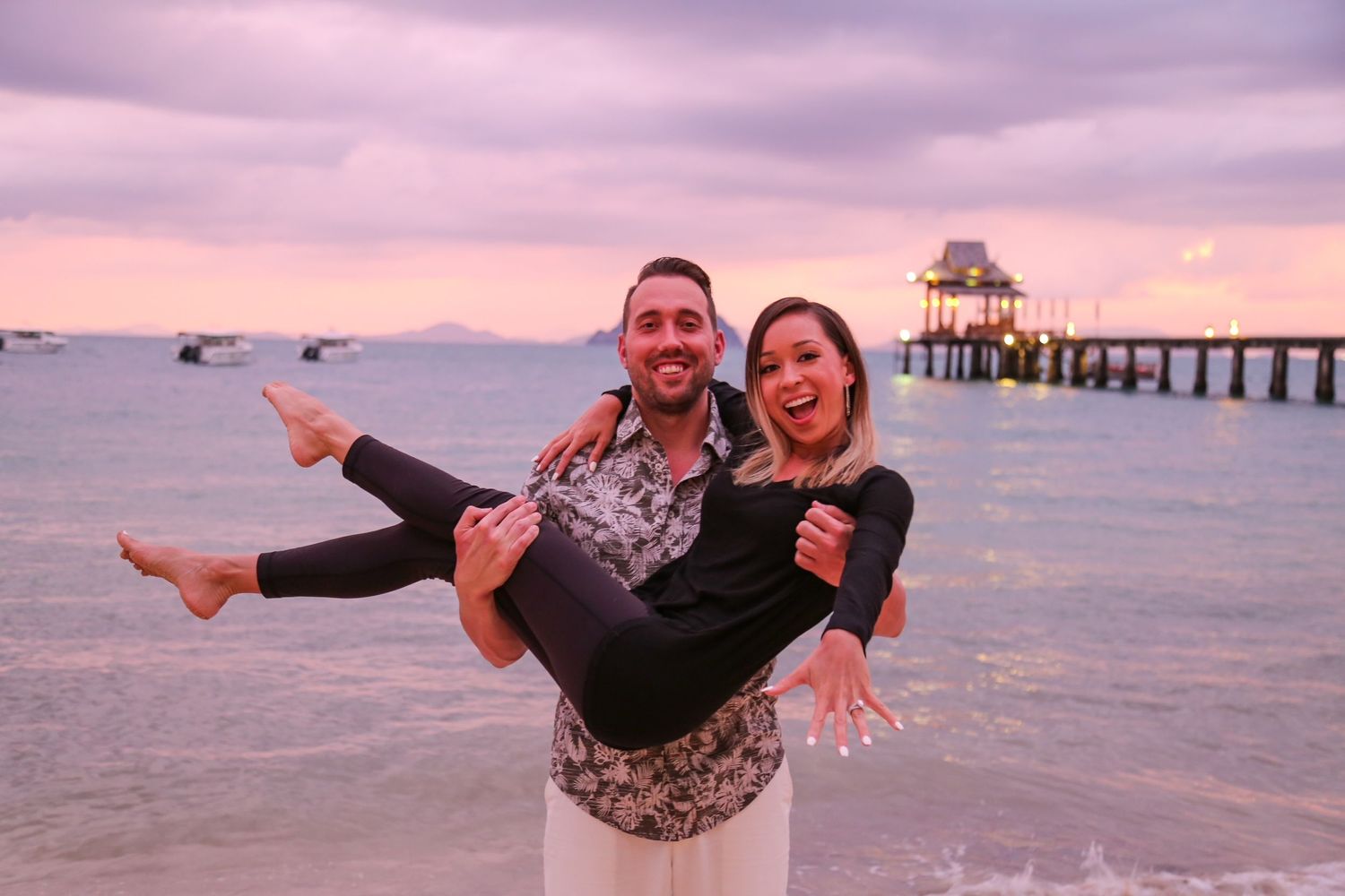 man holding wife as they pose on the beach in Thailand after they got engaged.