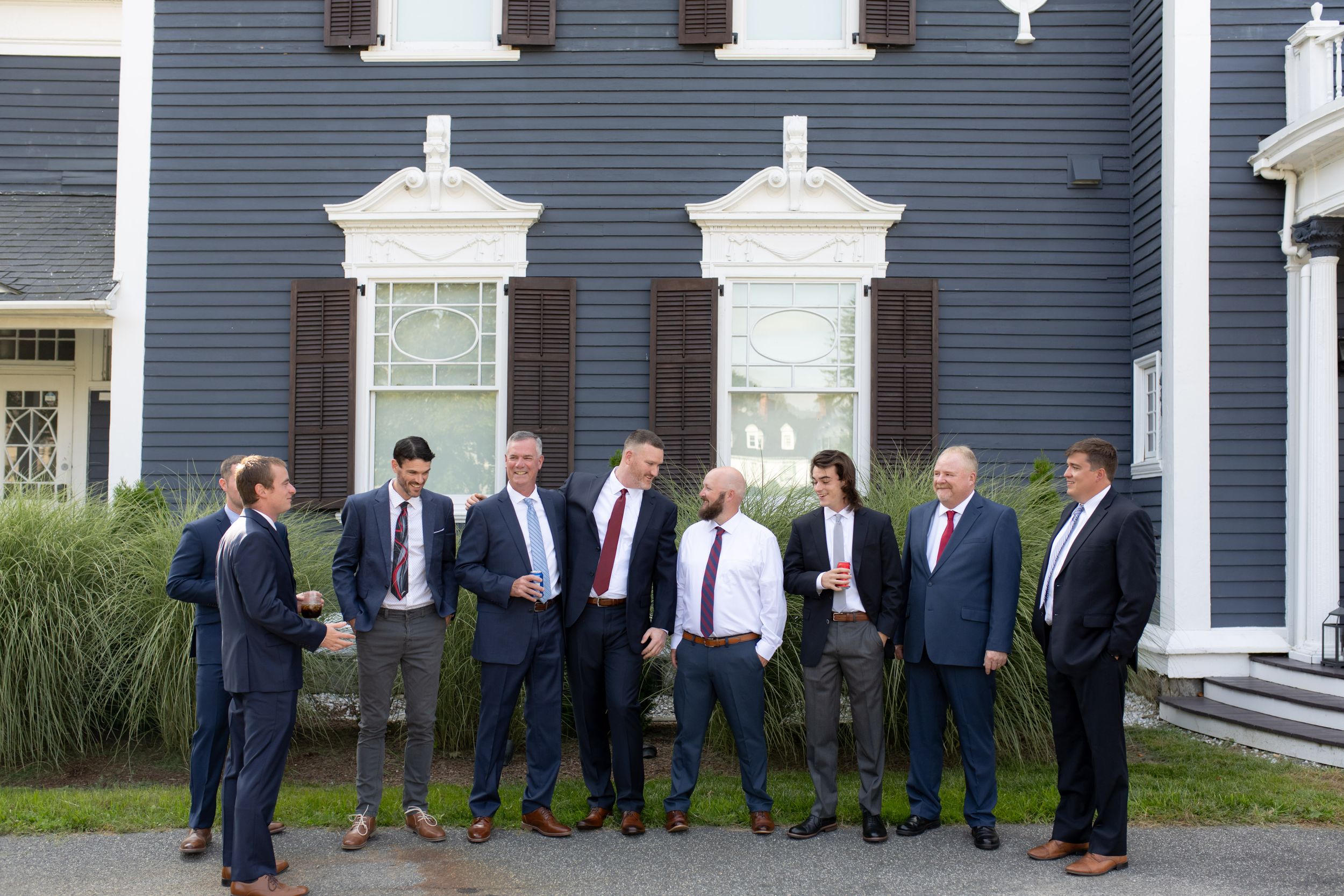 A group of formally dressed people stand in front of a classic navy blue mansion for a group photo.