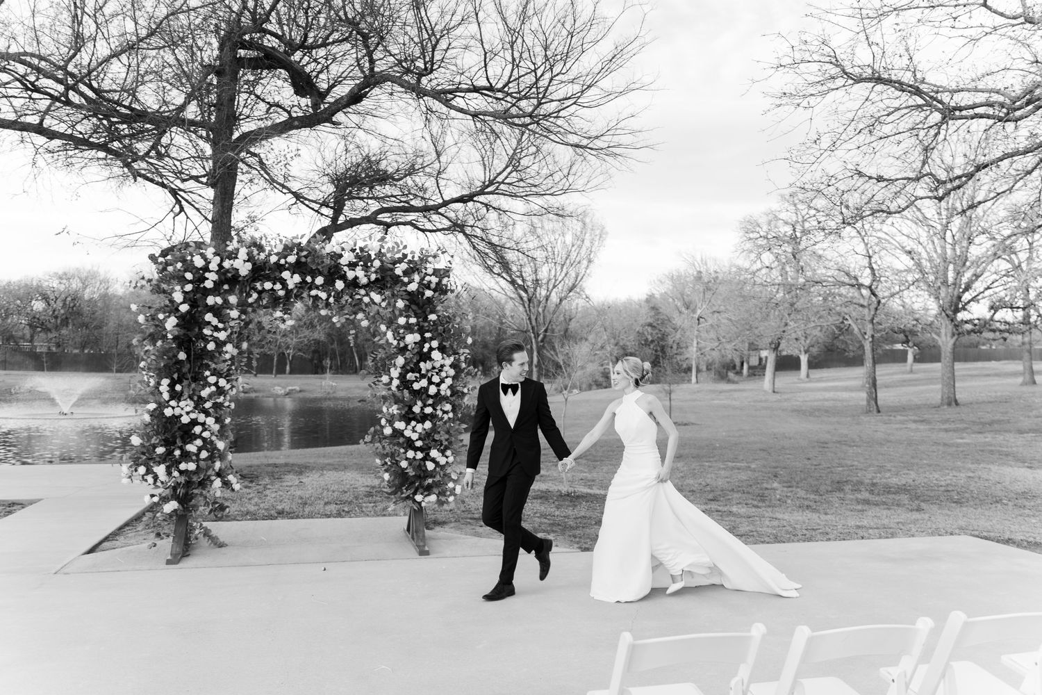 A black and white photo of a wedding ceremony setup with a floral arch and pond in the background.