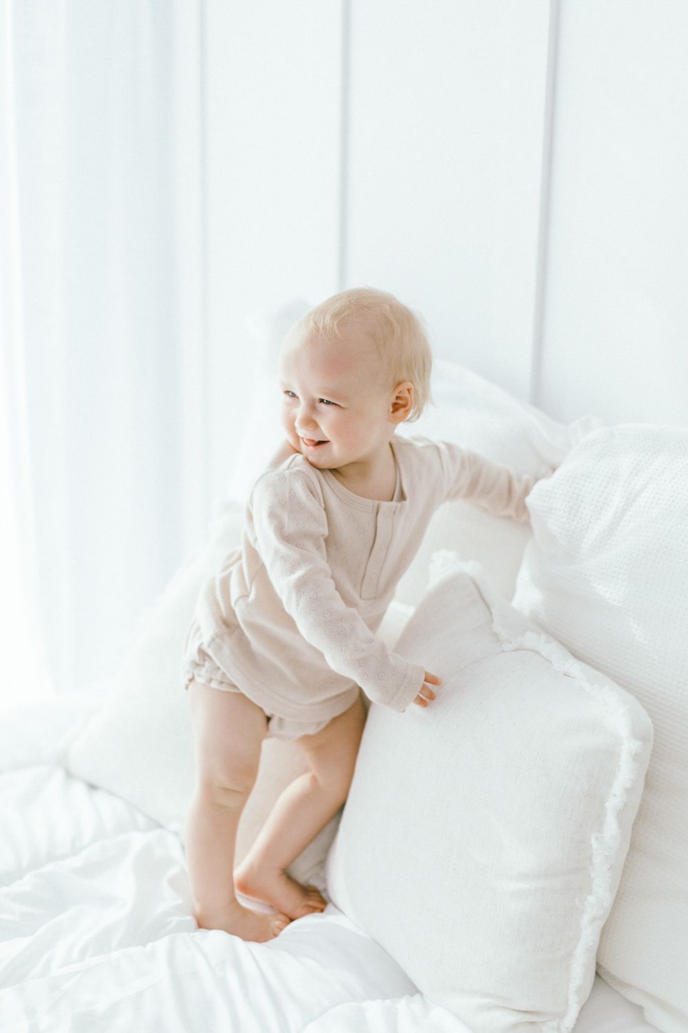 Baby in beige romper playing on white pillows with natural lighting.