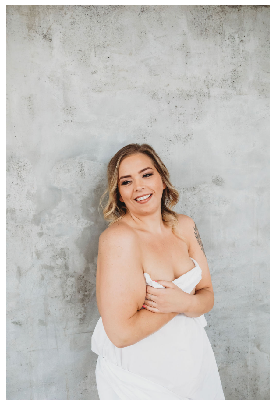 Woman wrapped in white sheet against concrete wall in artistic portrait session.