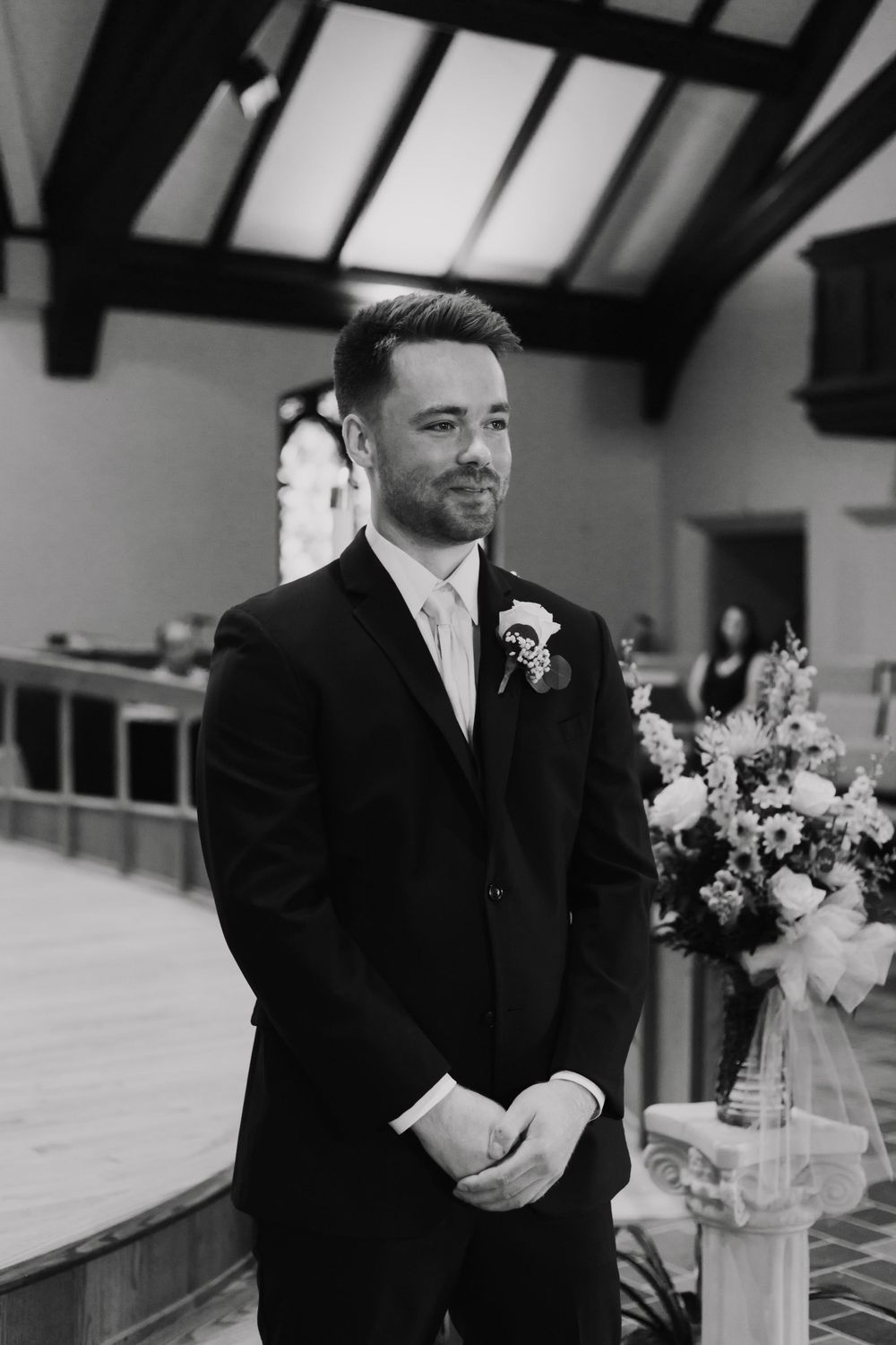 Groom in classic black suit with boutonniere standing in church interior.