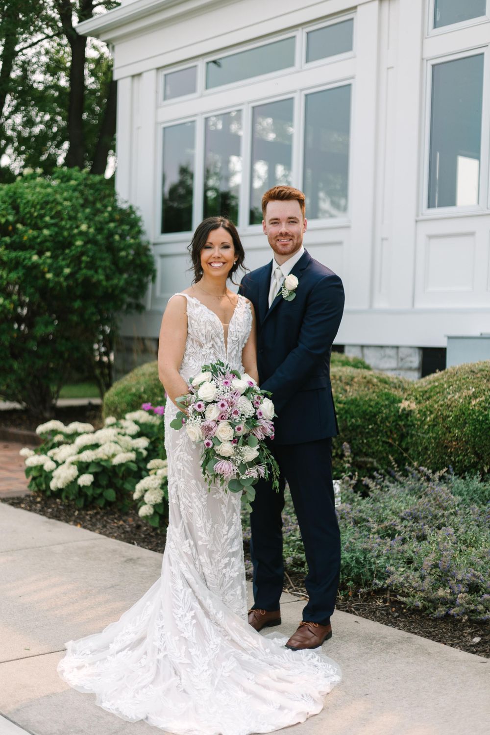 A wedding couple poses in front of a white building with elegant landscaping and hydrangeas.
