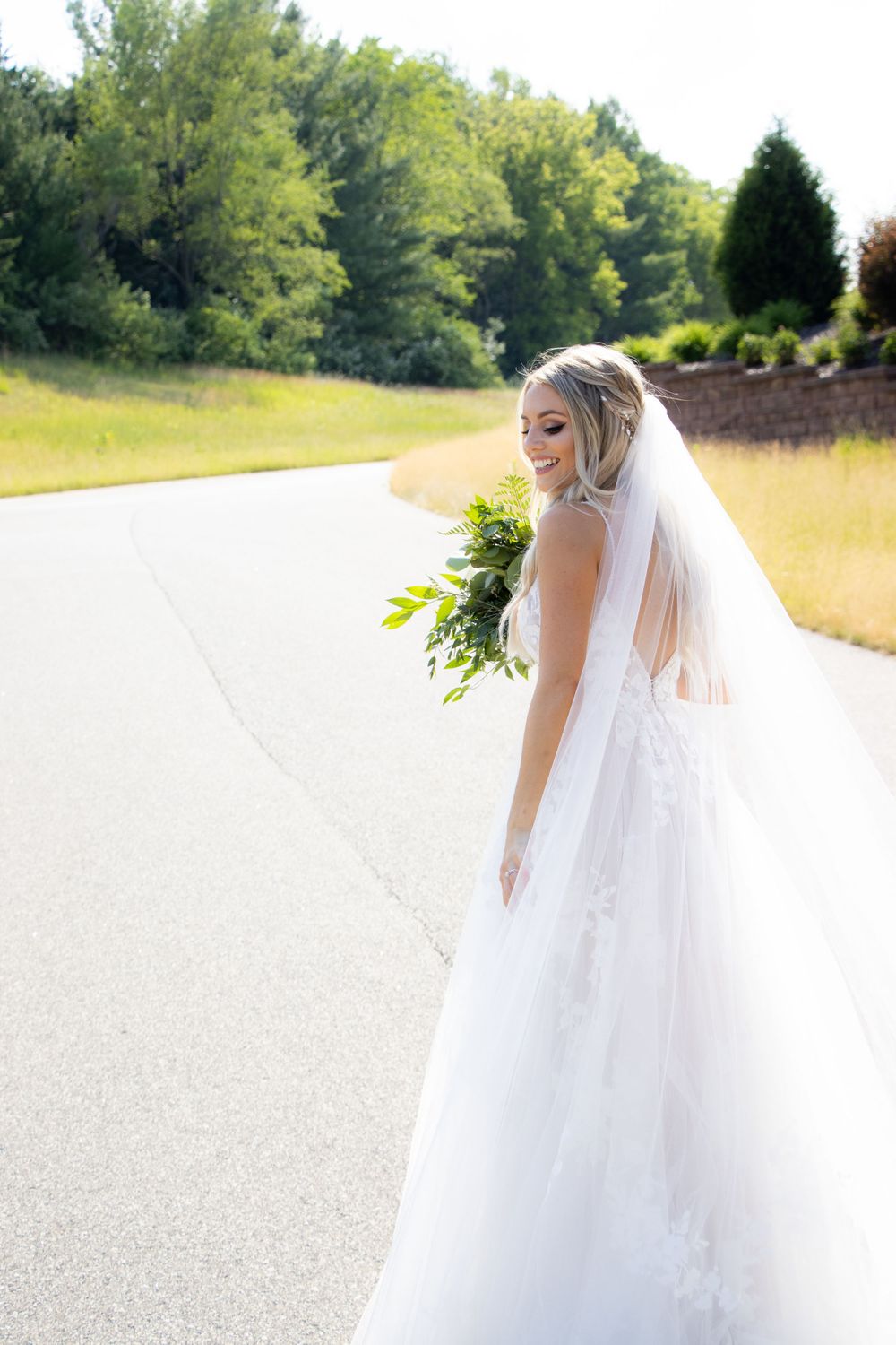 A bride in a white lace gown and veil stands on a paved pathway holding a greenery bouquet.