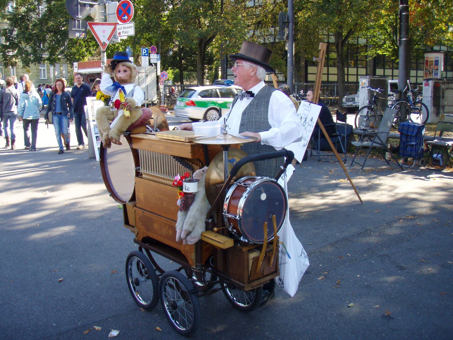 A traditional organ grinder performs with a barrel organ on wheels at an outdoor gathering in a park on a sunny day.