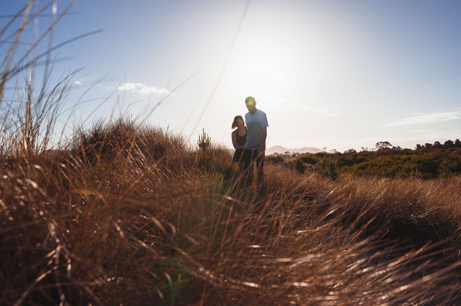 Couple standing in tall grass field backlit by bright sun on hilltop.