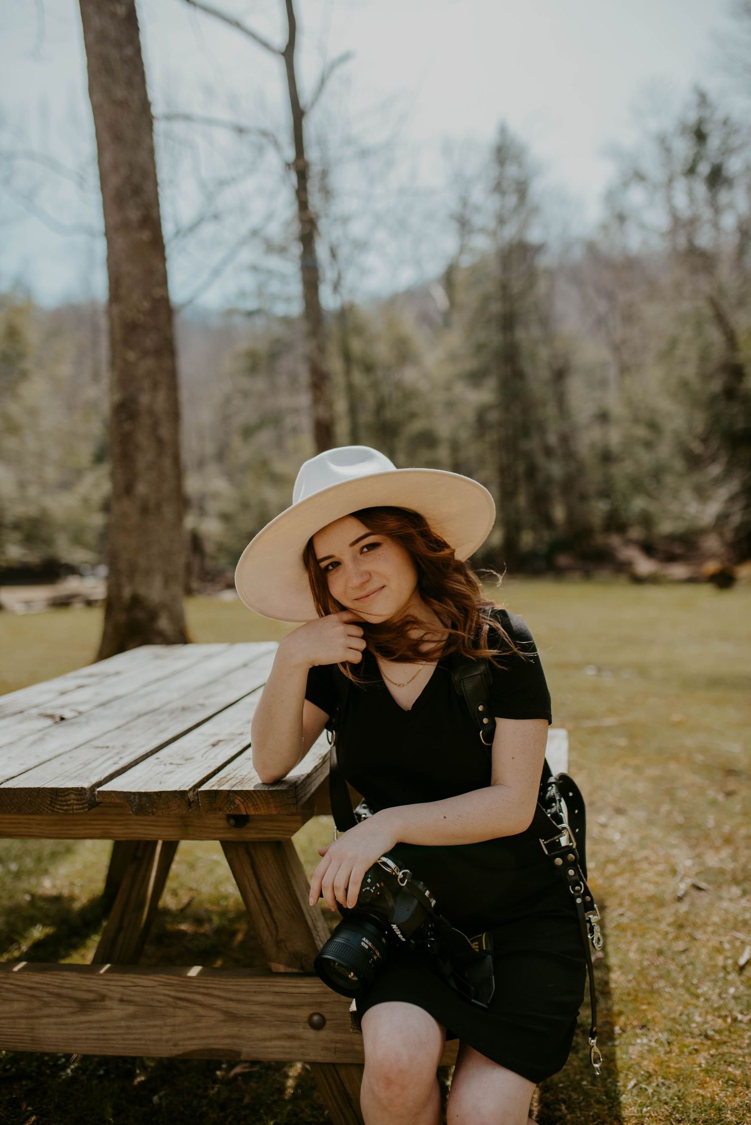 Person in black dress and white hat sitting at rustic picnic table in wooded setting.