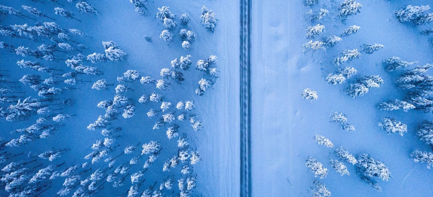 Birds-eye view of a straight road cutting through a dense snow-covered winter forest landscape.