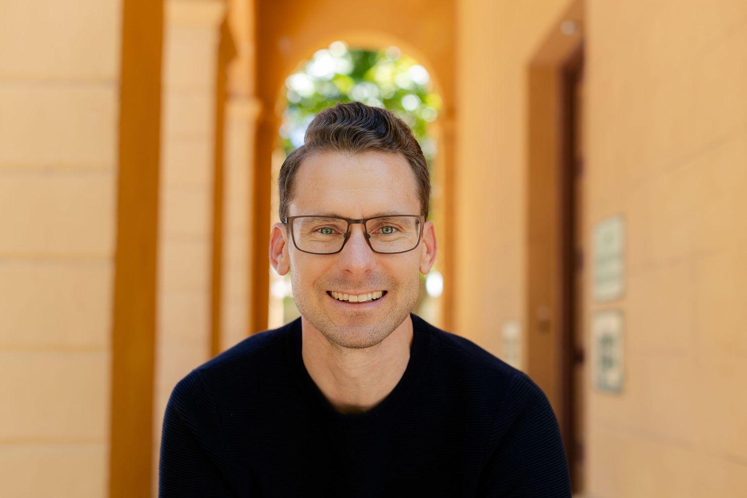 Professional headshot of person in black sweater smiling warmly against orange background.