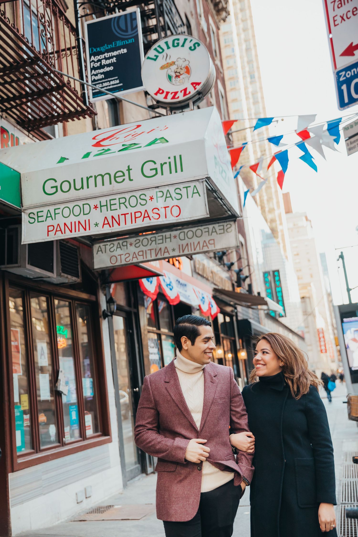 Smiling couple walking arm in arm past Luigi's Pizza and Gourmet Grill on a New York City street.