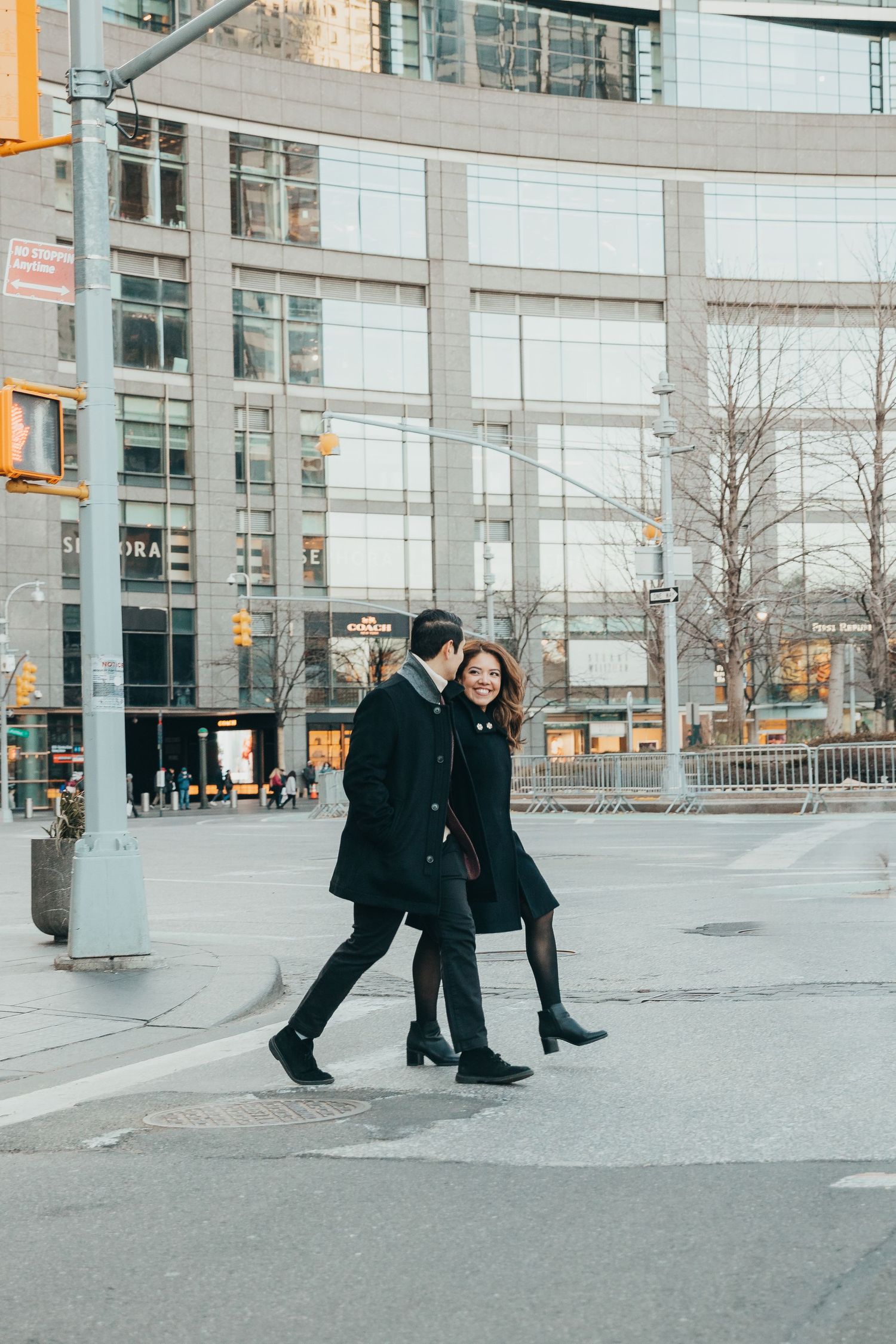 Couple in matching black coats walking and laughing together across a New York City crosswalk.