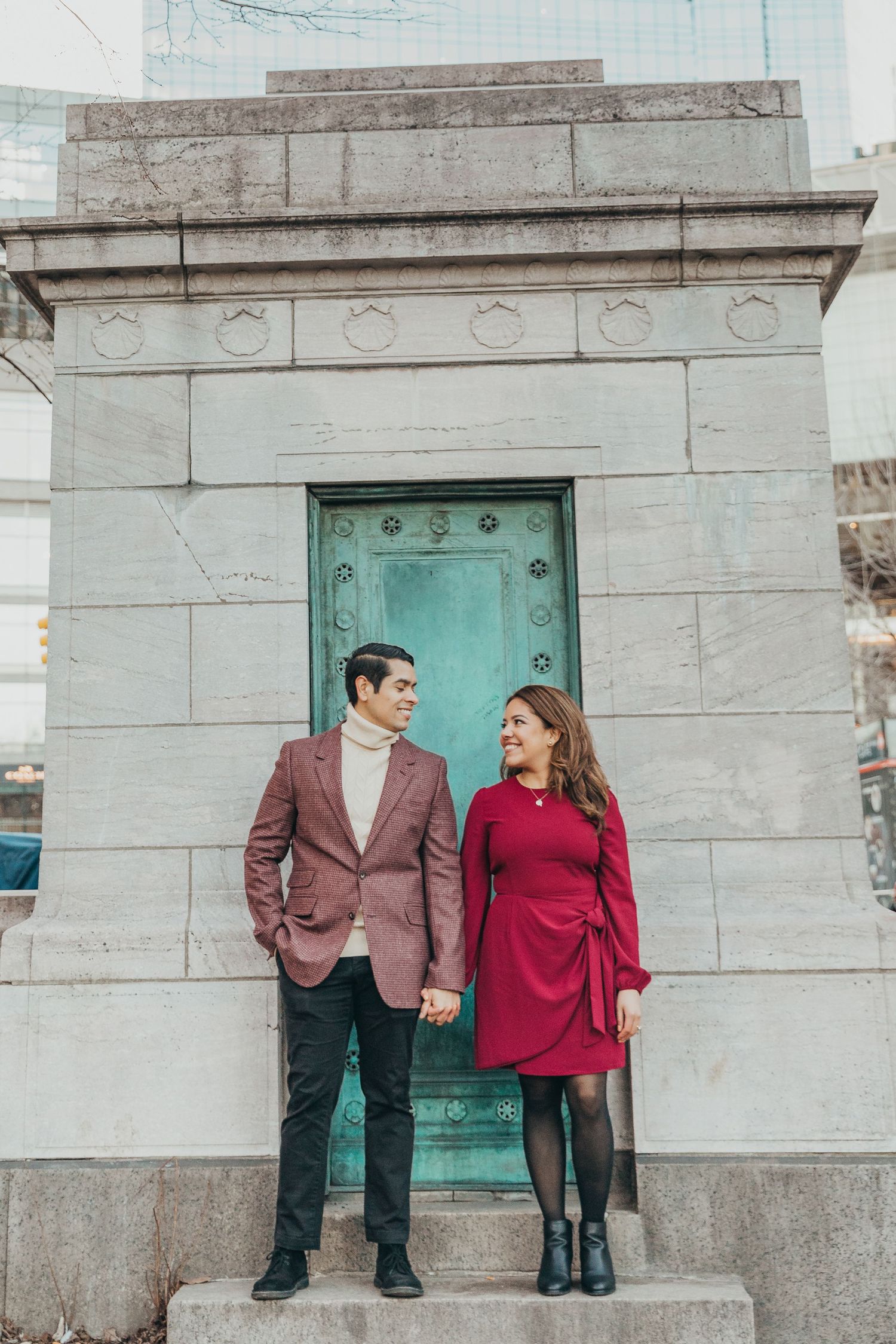 Couple holding hands and smiling at each other in front of a stone monument with bronze door.