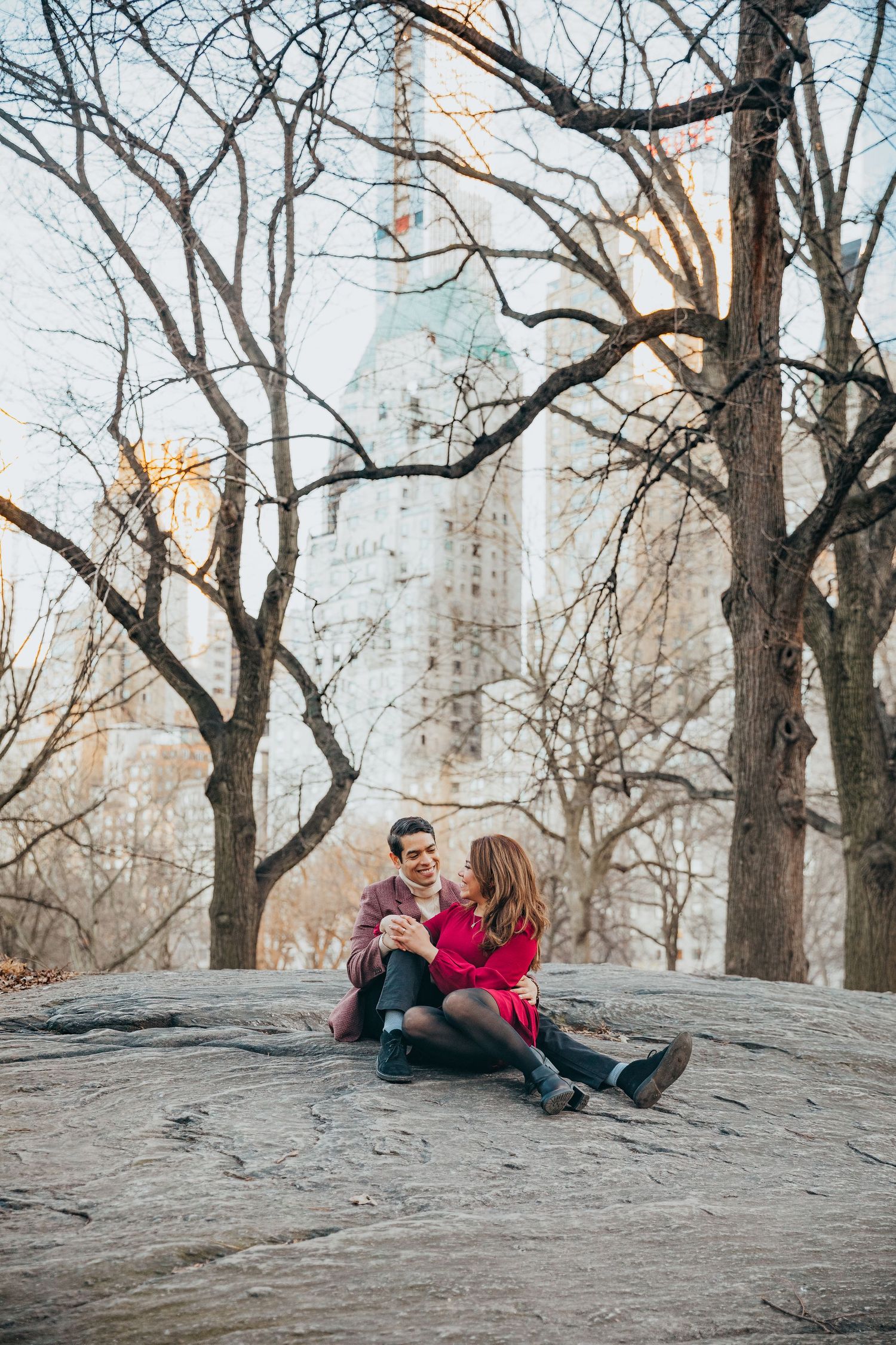 Couple sitting on rocky outcrop in Central Park with The Plaza Hotel visible through bare trees.