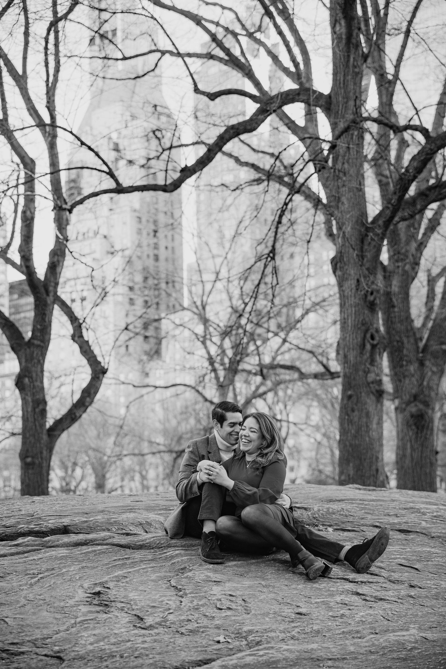 Black and white photo of couple sitting on rock in Central Park with NYC skyline behind them.