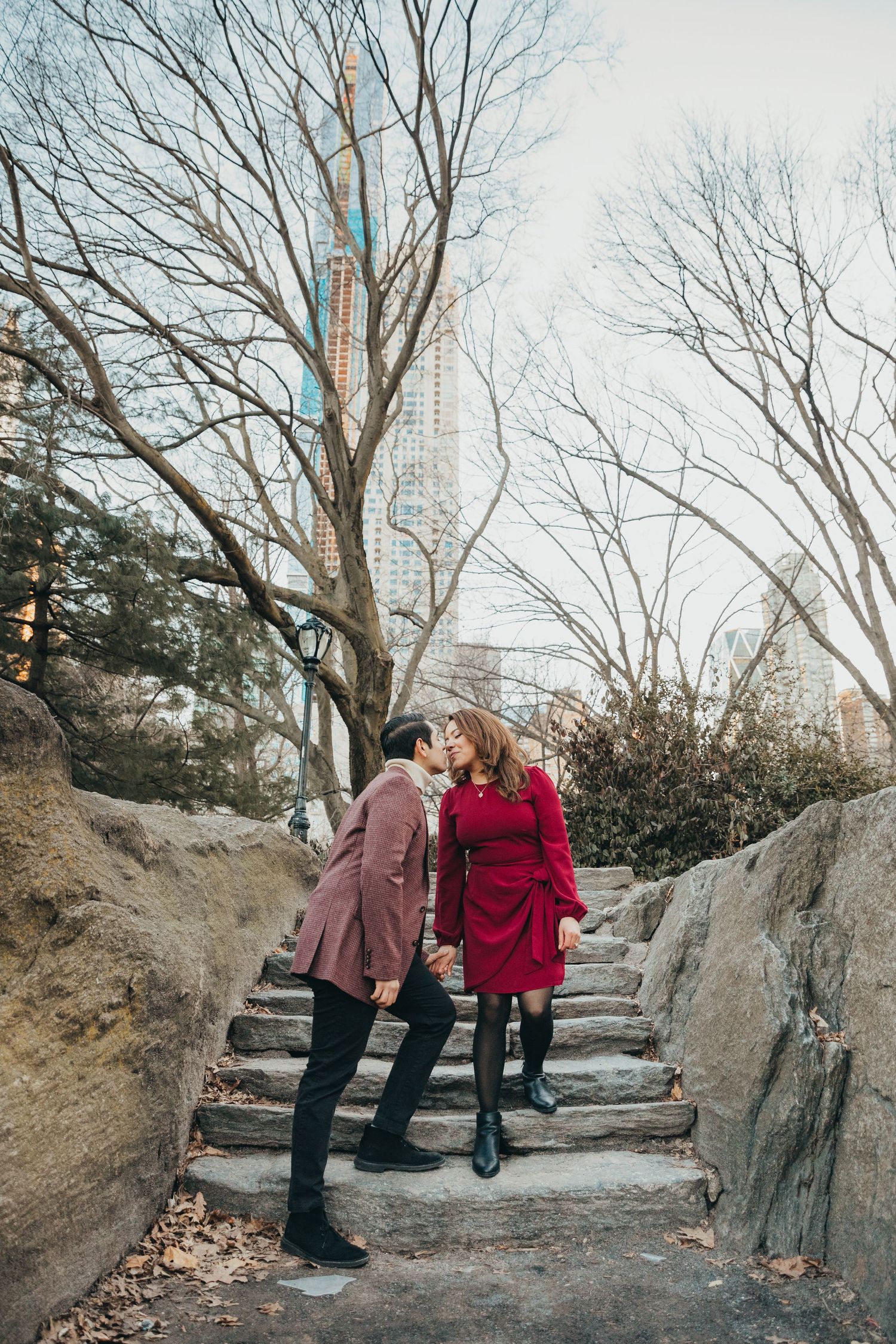Couple sharing a kiss on stone steps in Central Park with city skyscrapers visible through bare winter trees.