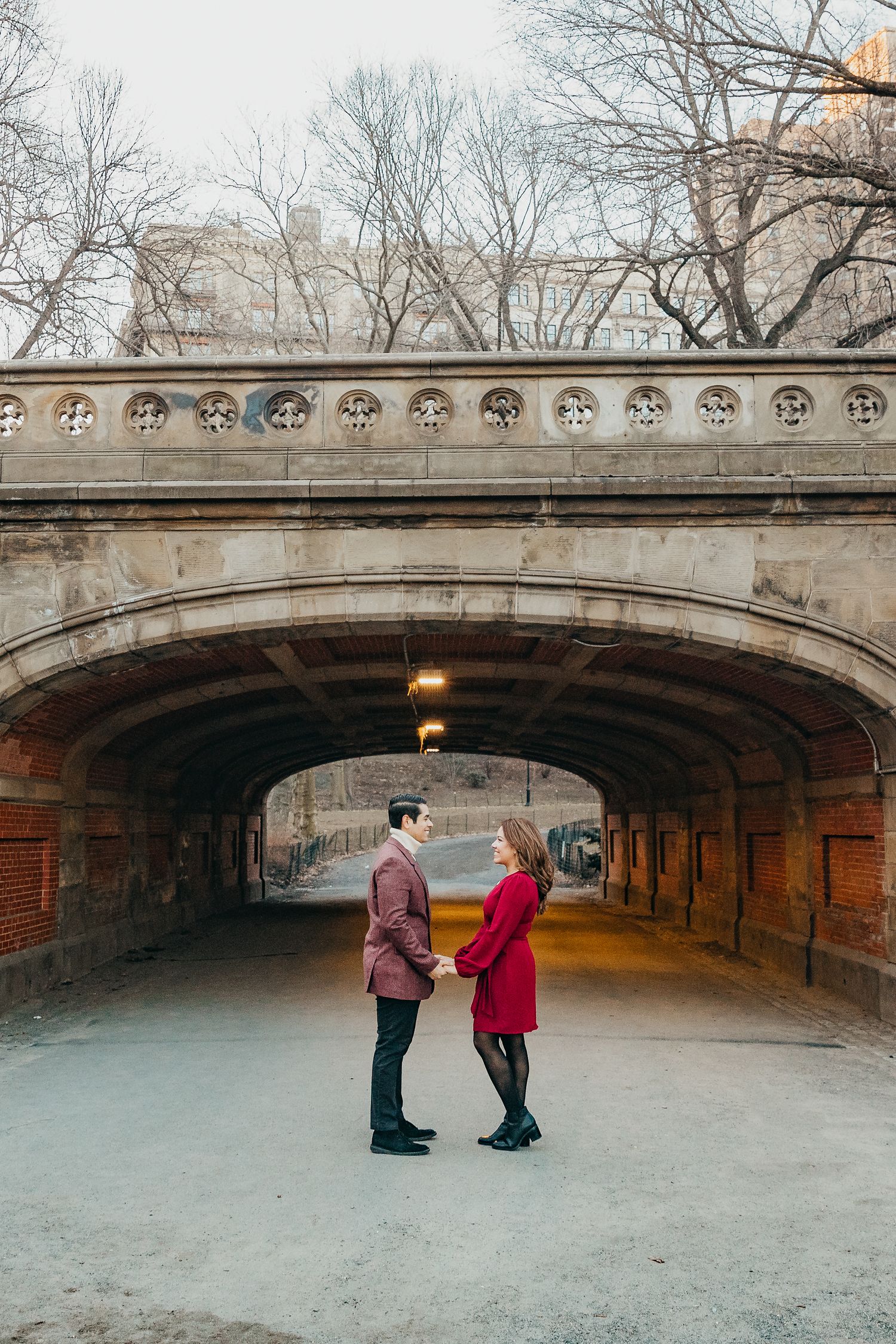 Couple holding hands and facing each other beneath a stone arch bridge in Central Park during winter engagement.