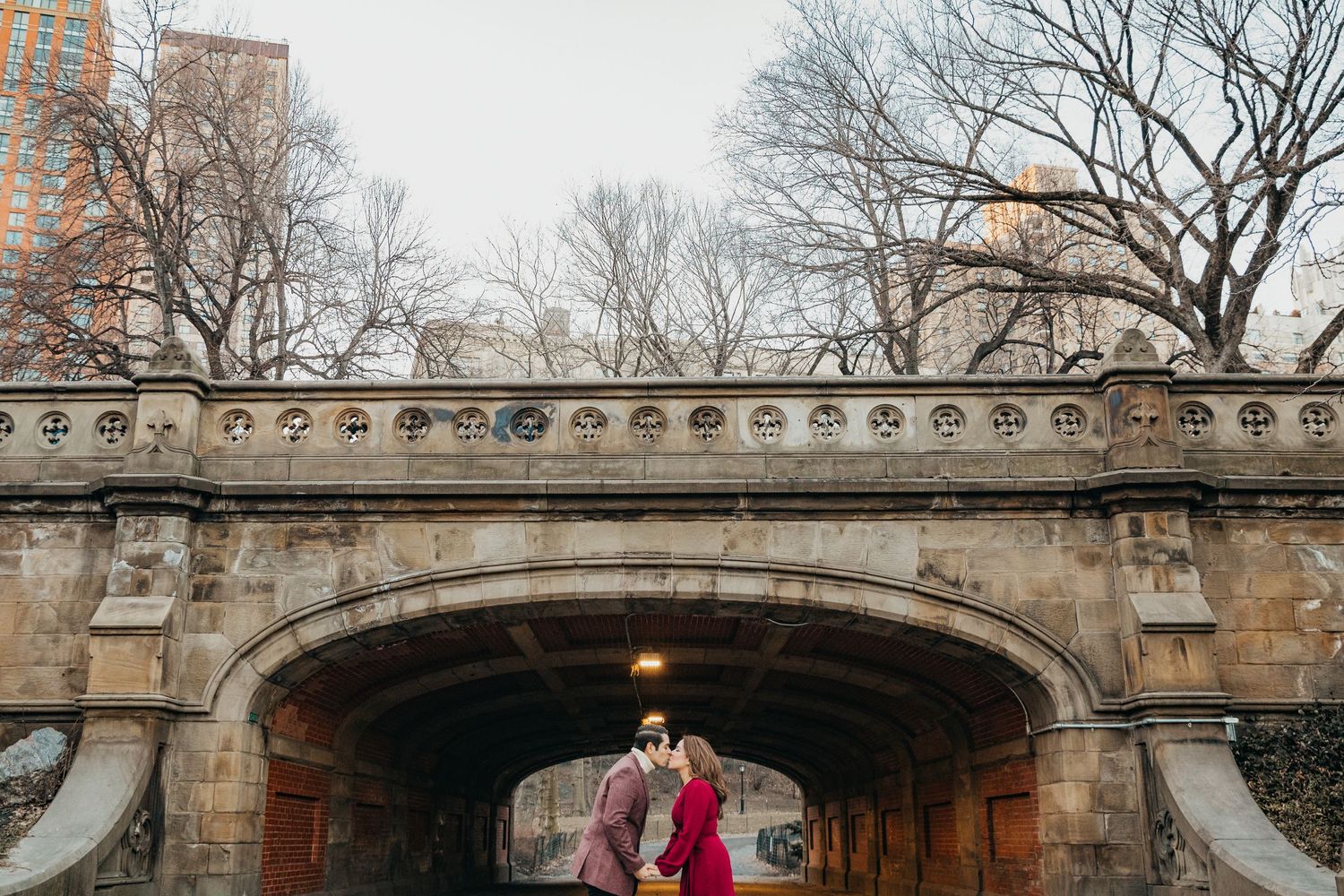 Couple kissing beneath a grand stone arch bridge in Central Park during a romantic winter engagement session.