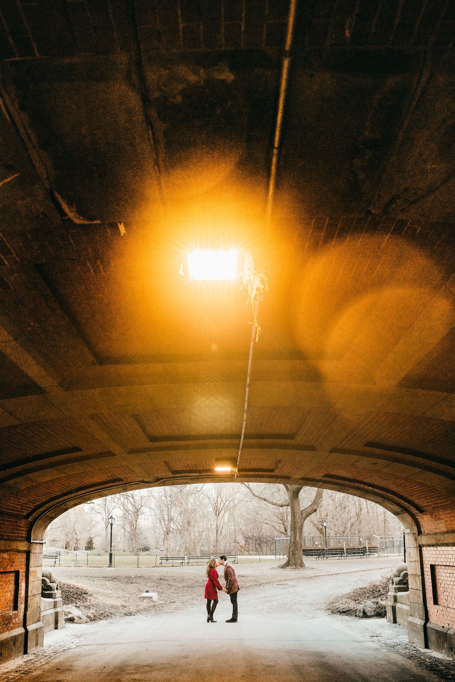 Couple holding hands under a glowing bridge tunnel in a snowy Central Park during a winter engagement session.