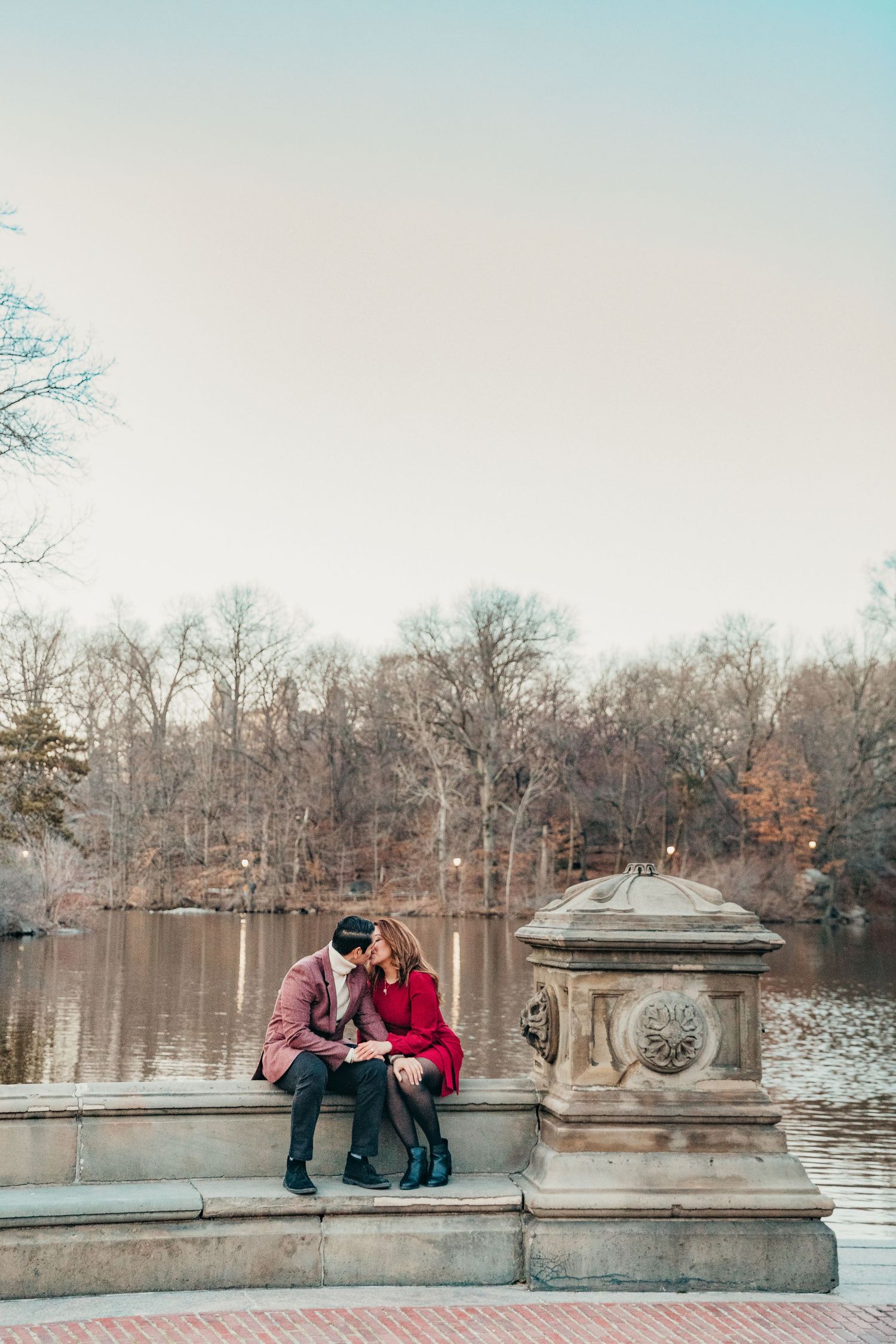 Romantic couple sitting close together on stone steps by a lake in Central Park during autumn.