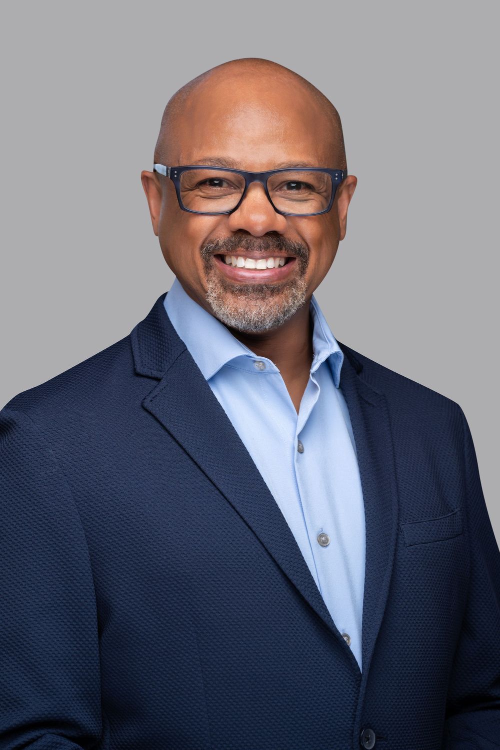 Professional headshot of person in navy blazer and light blue shirt against grey background.