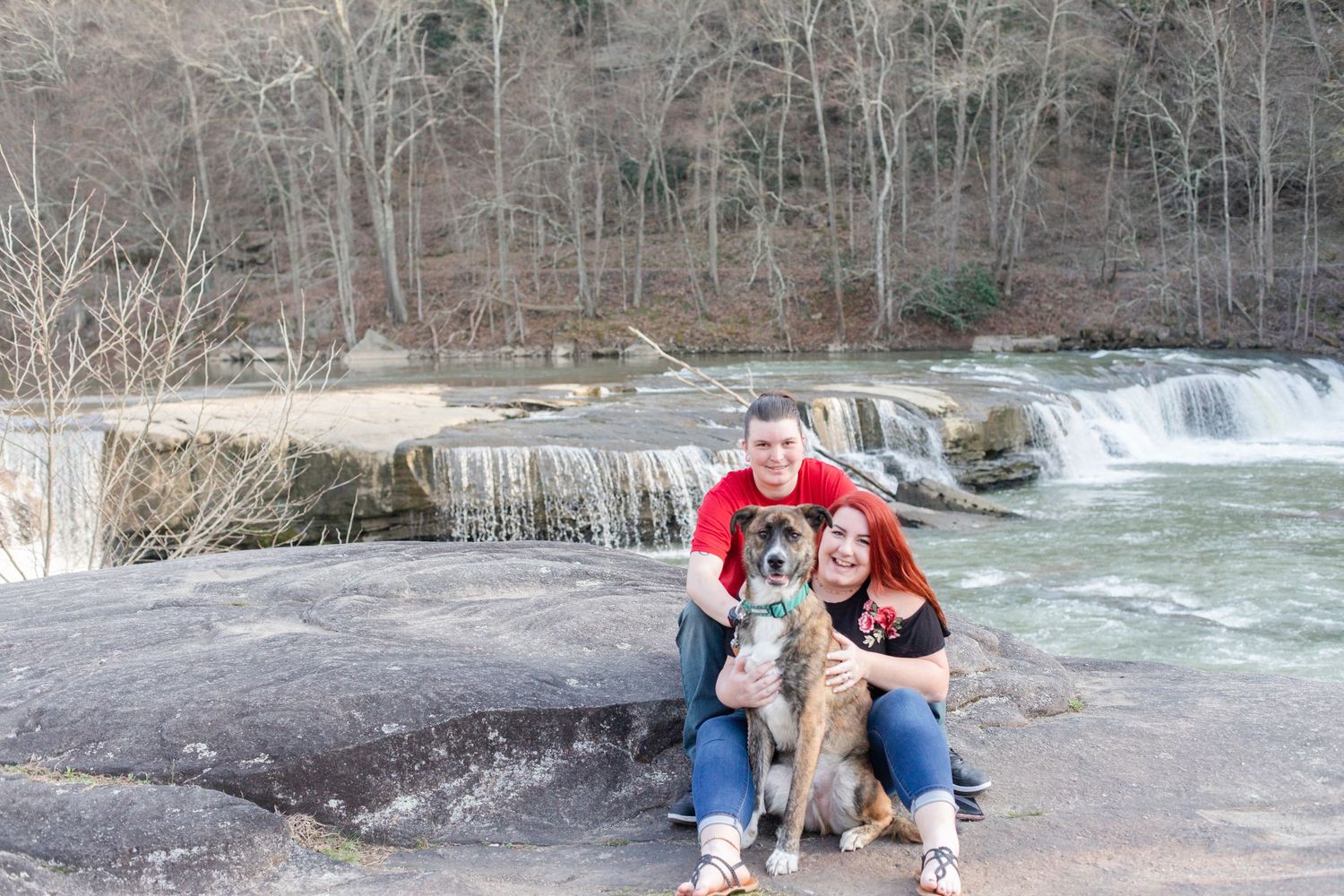A couple sits with their dog on rocky terrain near a flowing waterfall.