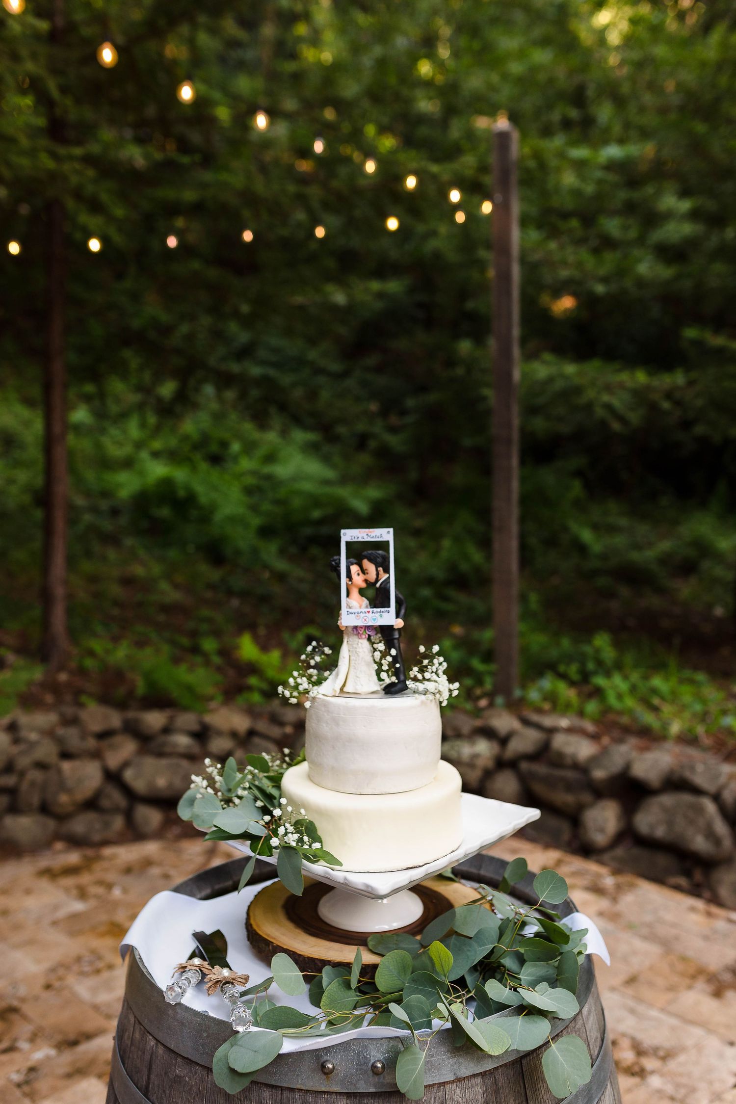 White wedding cake decorated with eucalyptus leaves sits on a wine barrel under string lights.