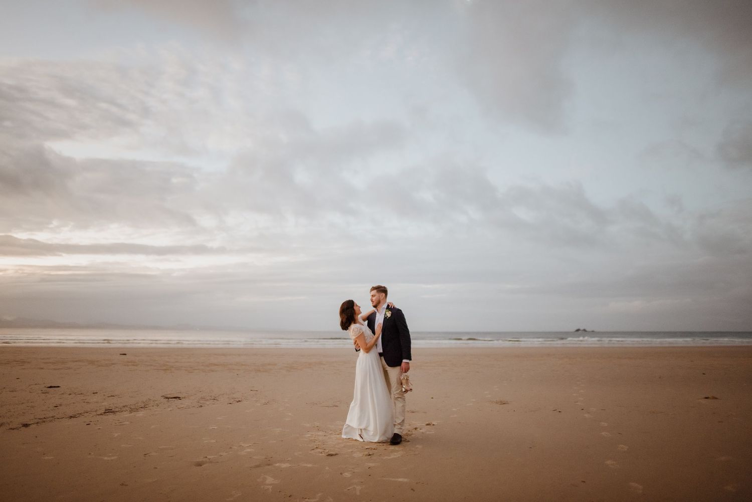 Bride and groom stand together on a sandy beach under a cloudy sky.