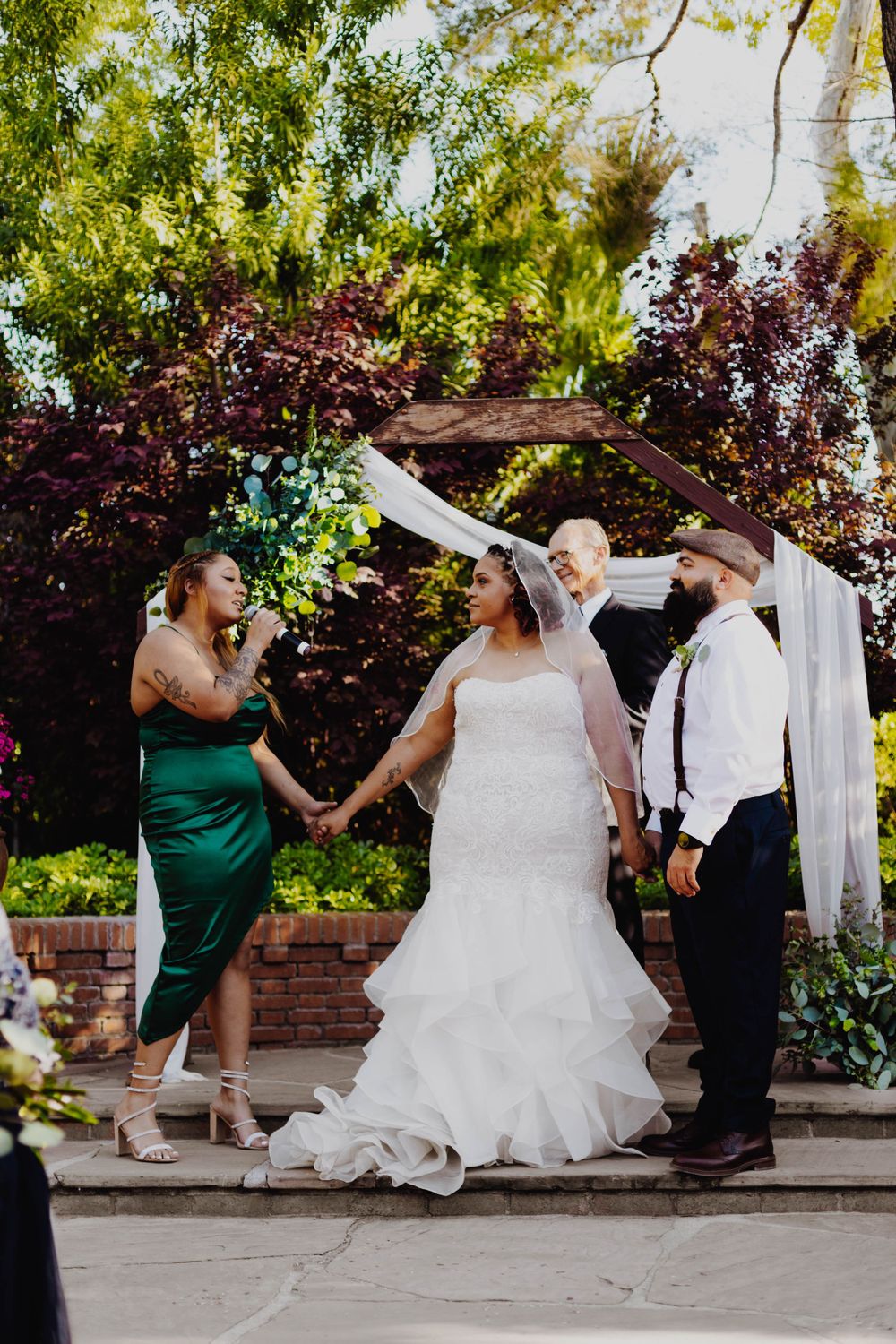 Wedding party interacts on garden steps with decorative white fabric arch overhead.