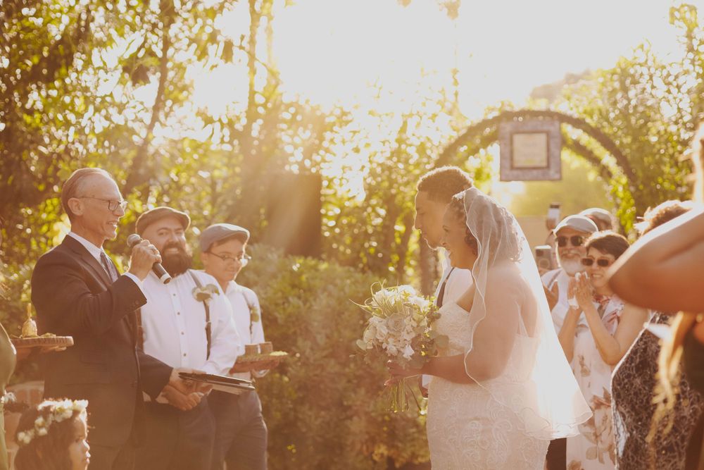 Golden sunlight illuminates an outdoor wedding ceremony with guests gathered around a brick wall.