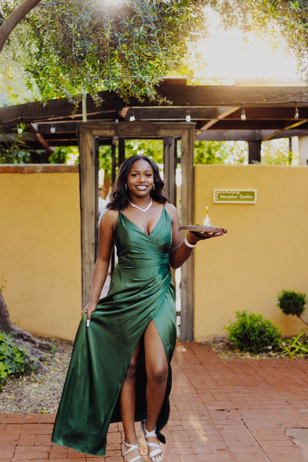 Elegant woman in emerald green formal dress poses in garden setting holding a drink.