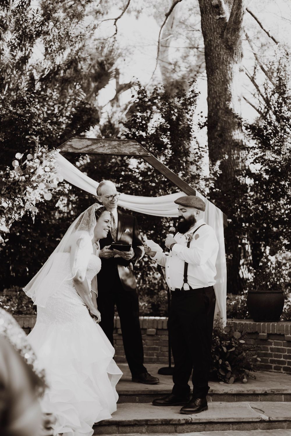 Black and white photo of wedding ceremony under draped fabric arch in garden setting.