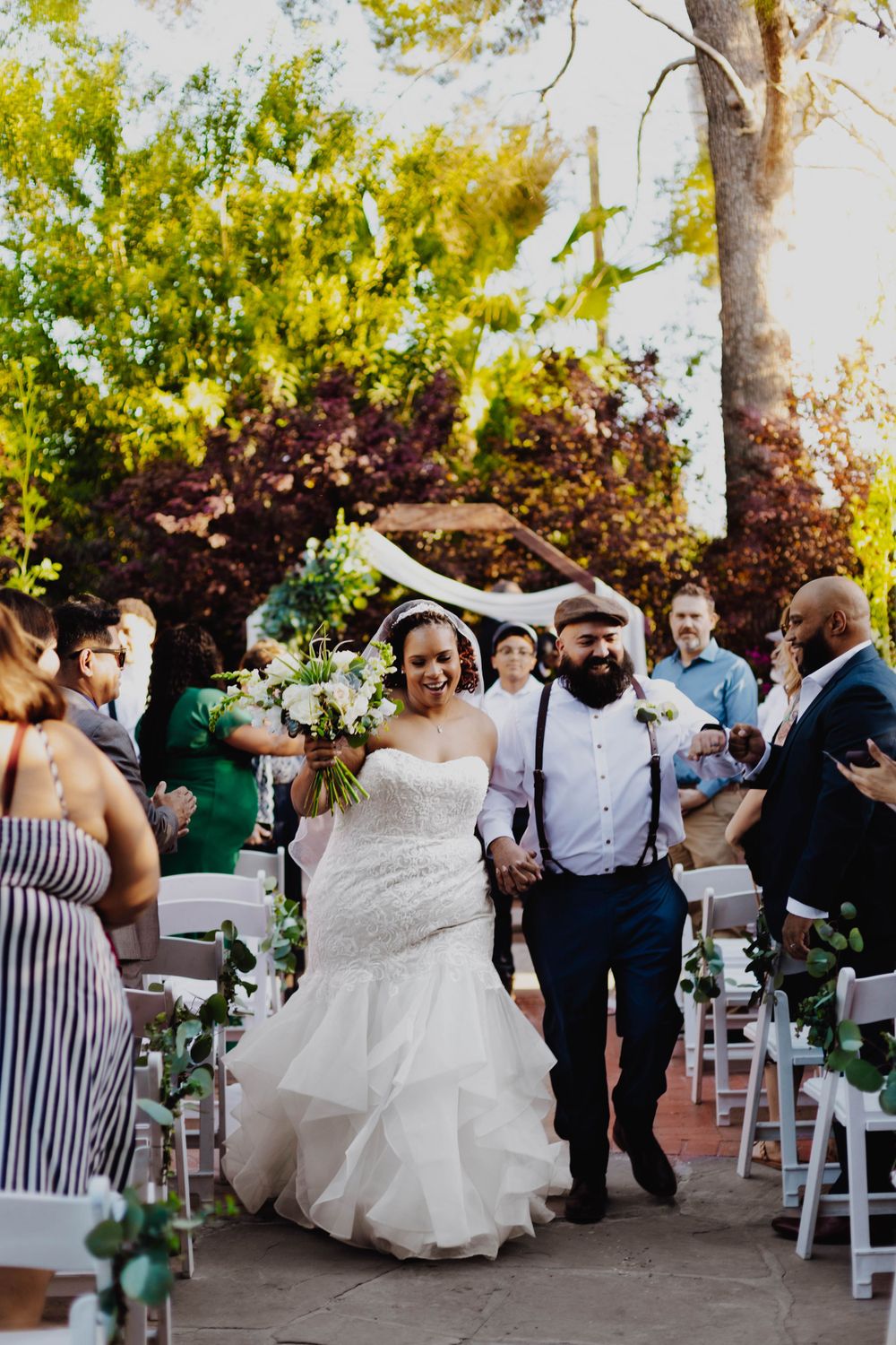 Wedding exit with guests celebrating in outdoor garden pathway lined with white chairs.
