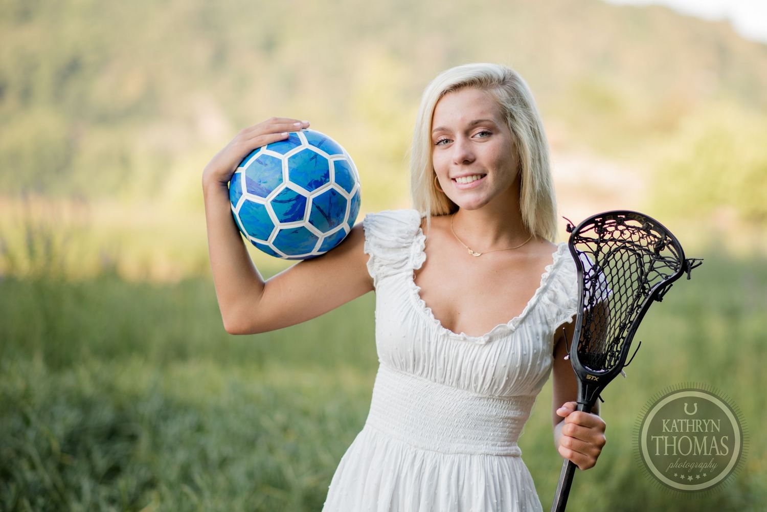 Woman in white dress poses with a soccer ball and lacrosse stick in a sunny field.