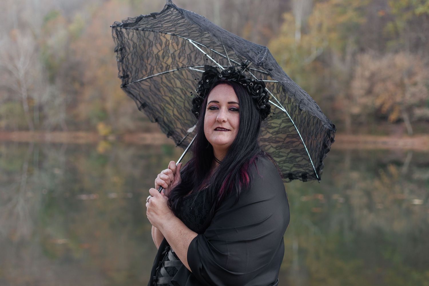 Gothic-styled figures with black parasols pose near a serene lake in autumn.