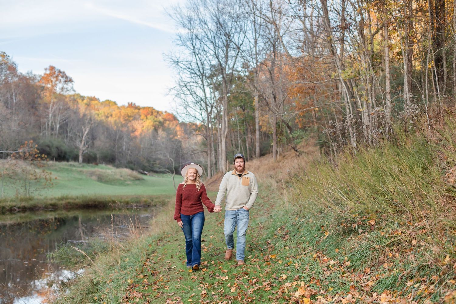 Scenic autumn engagement photos in a field with fall foliage and trees in the background.