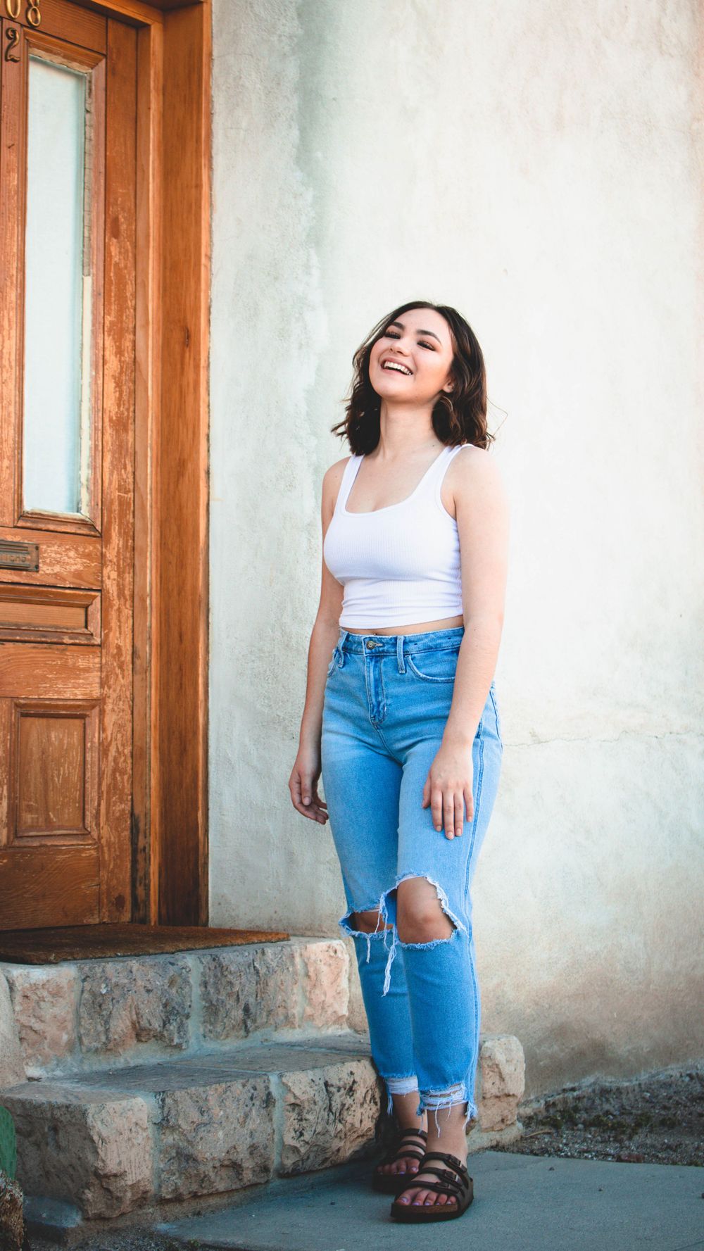 Person in white tank top and ripped jeans sits casually on stone steps by wooden door.