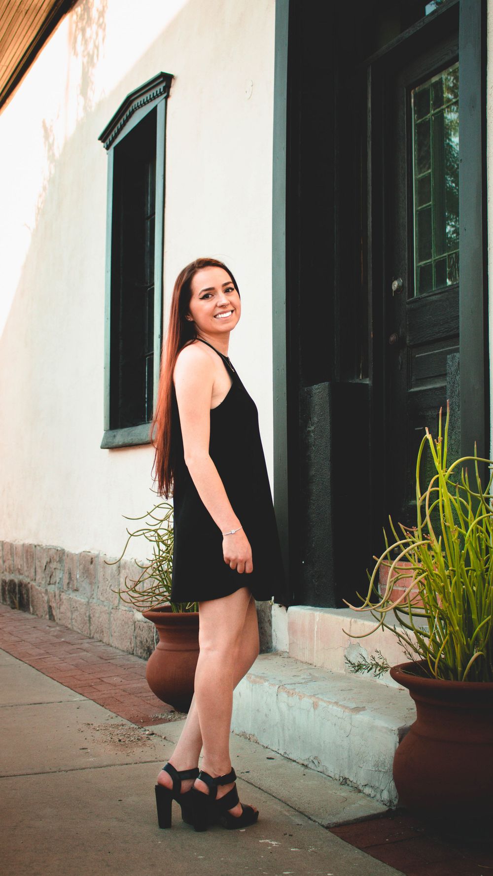 Woman in black dress poses beside doorway of historic building with plants.
