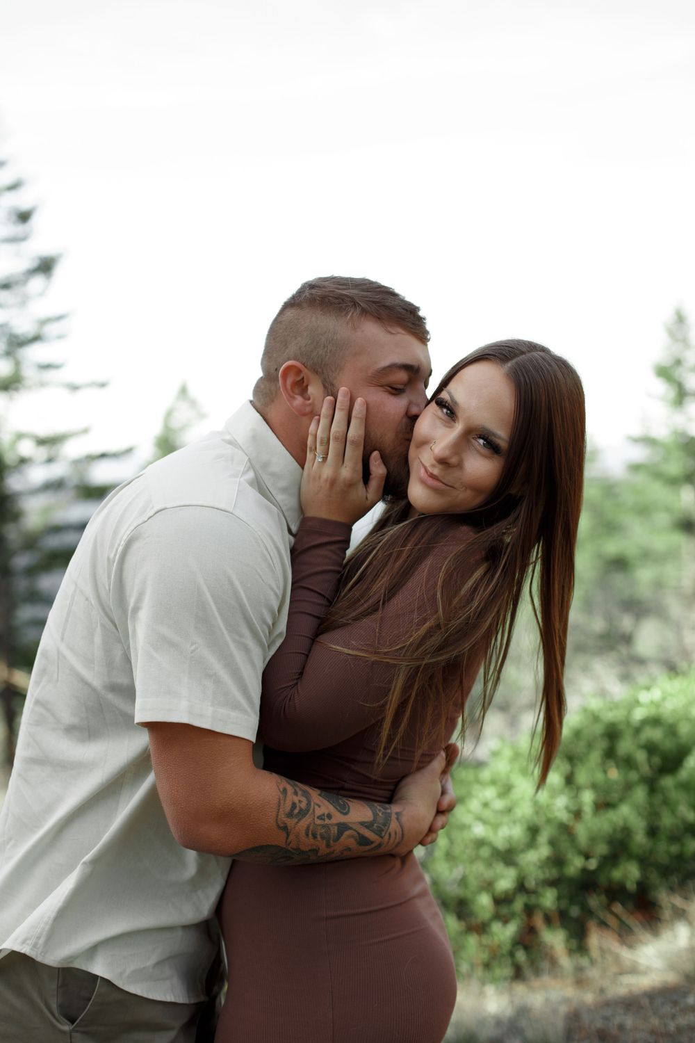 Romantic couple embraces outdoors in a natural setting with evergreen trees in the background.